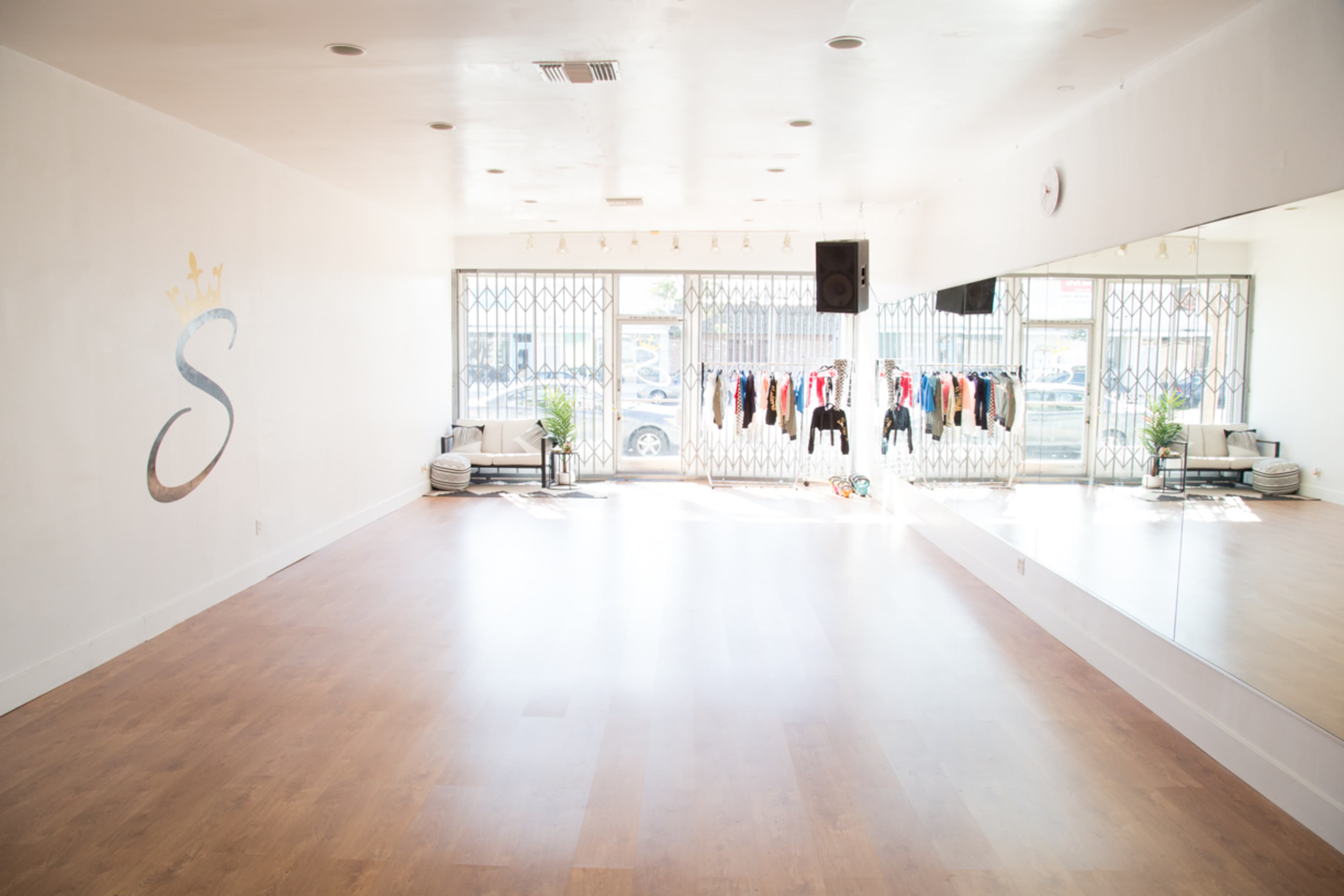 The image shows a spacious, empty dance studio featuring large windows, a mirrored wall, and a rack of clothing.