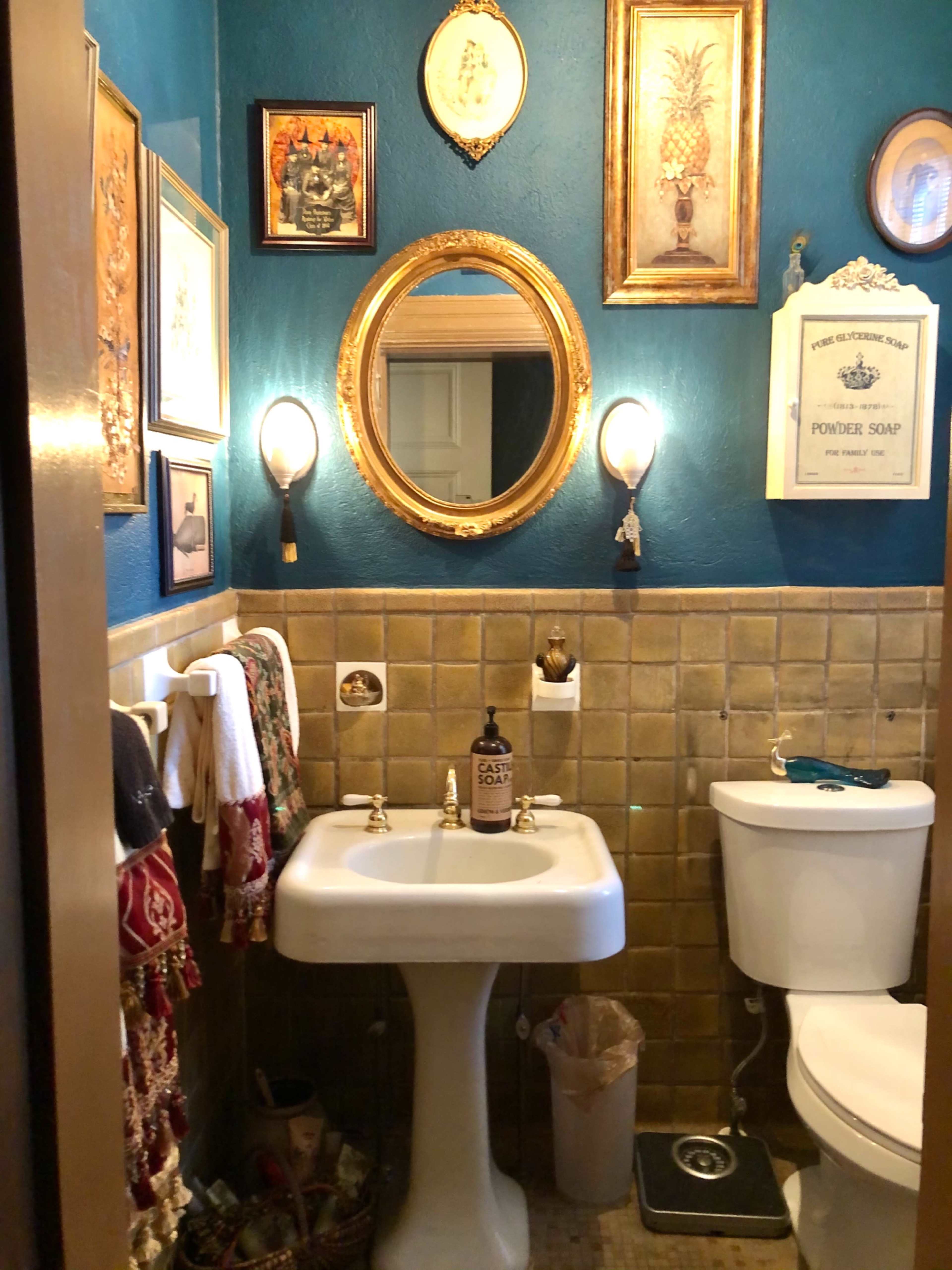 A small bathroom with a vintage sink, toilet, and various framed artworks on the teal walls.
