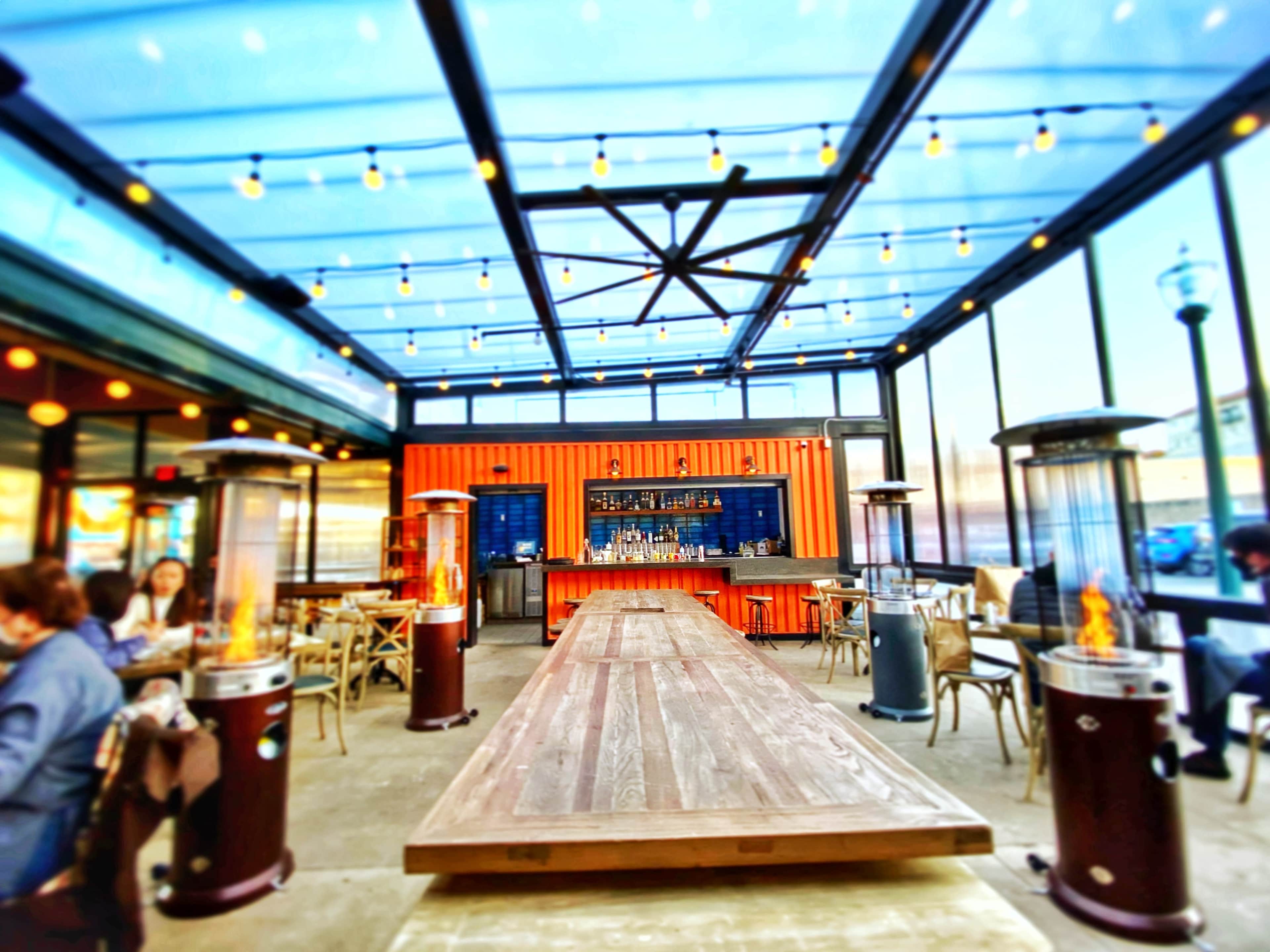 A modern indoor-outdoor restaurant with a long wooden table, glass walls, and decorative heating lamps.