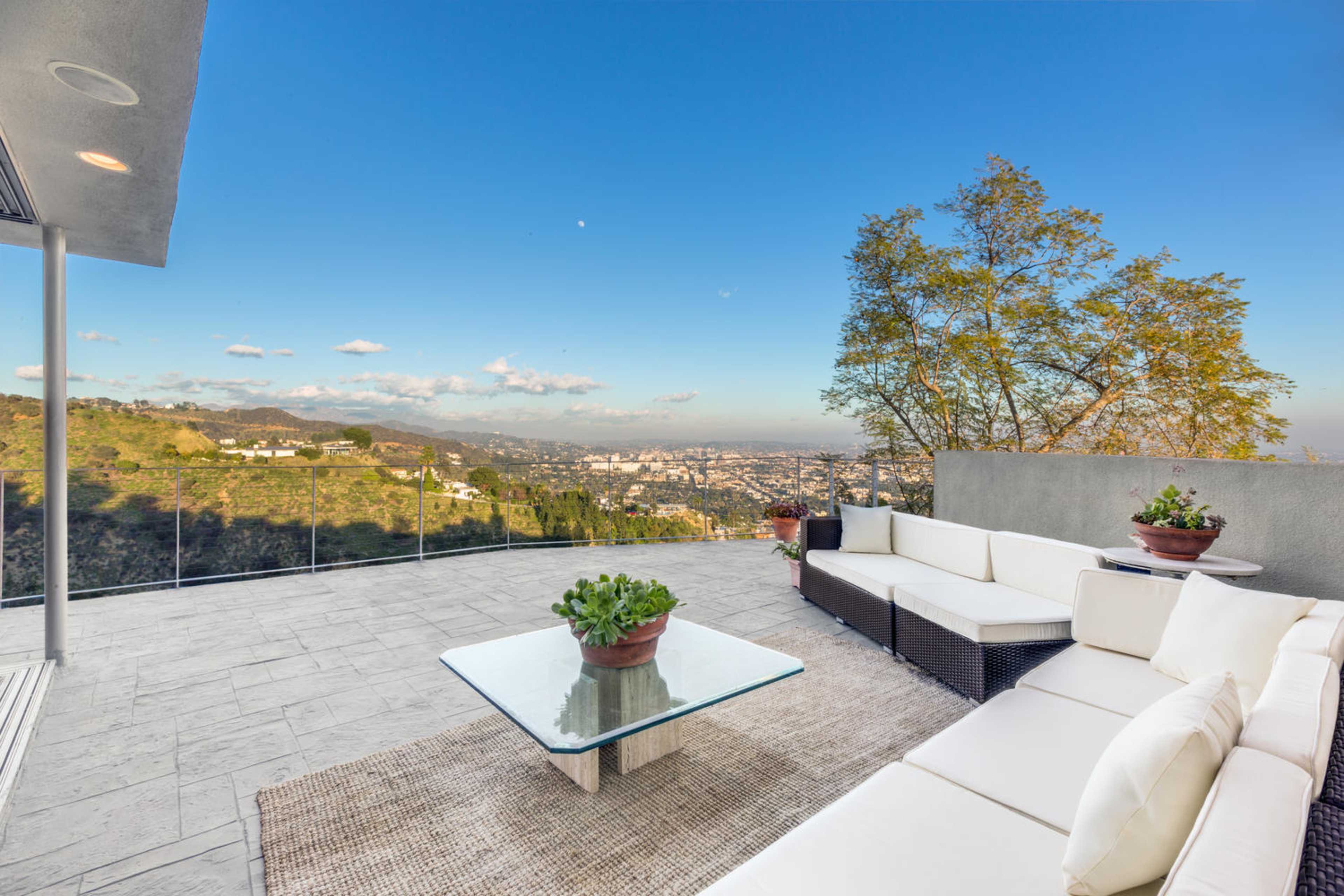 A modern outdoor patio with a glass table and white sofas, overlooking a city view and mountains in the background.