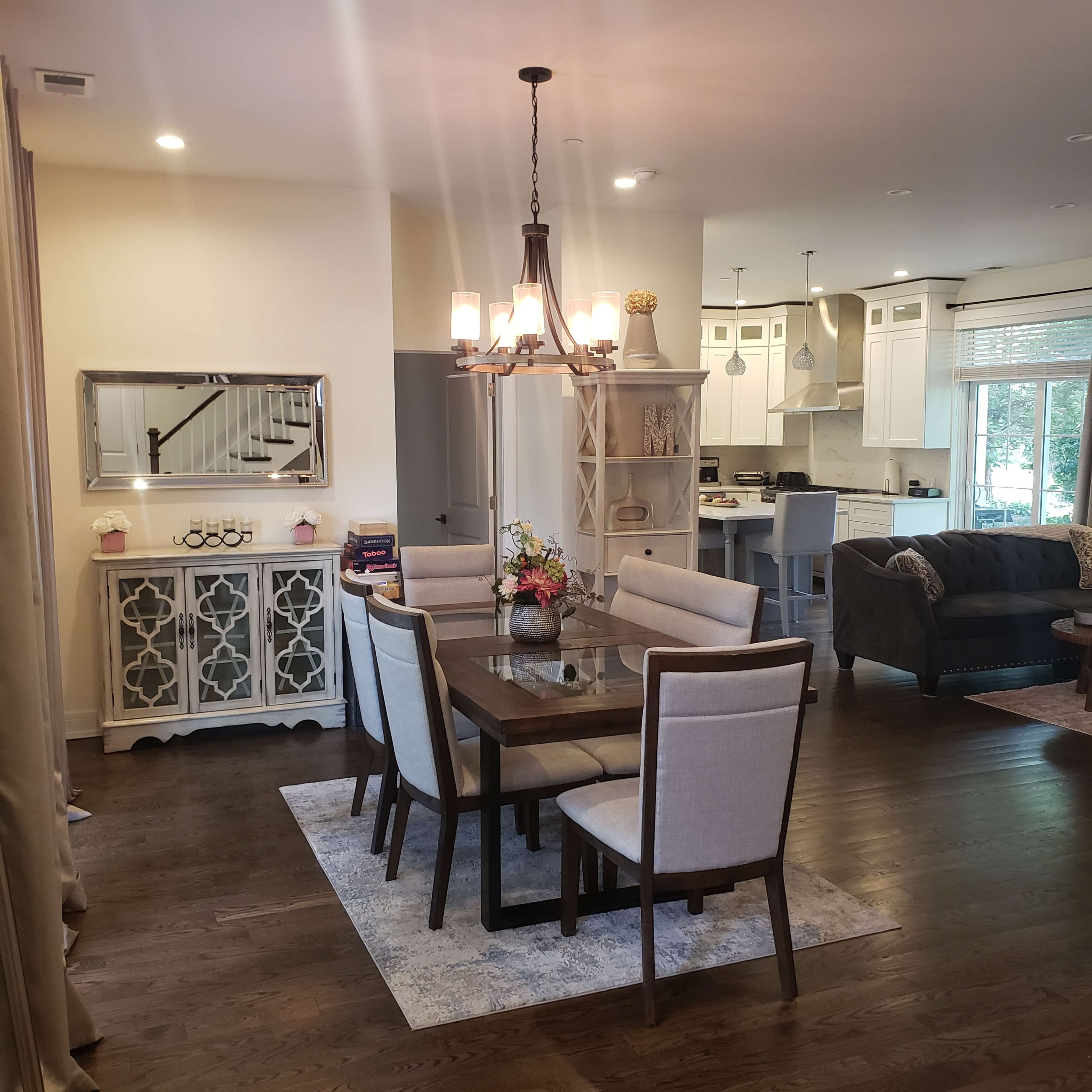A dining area features a wooden table surrounded by upholstered chairs, with a chandelier overhead and an open kitchen visible in the background.