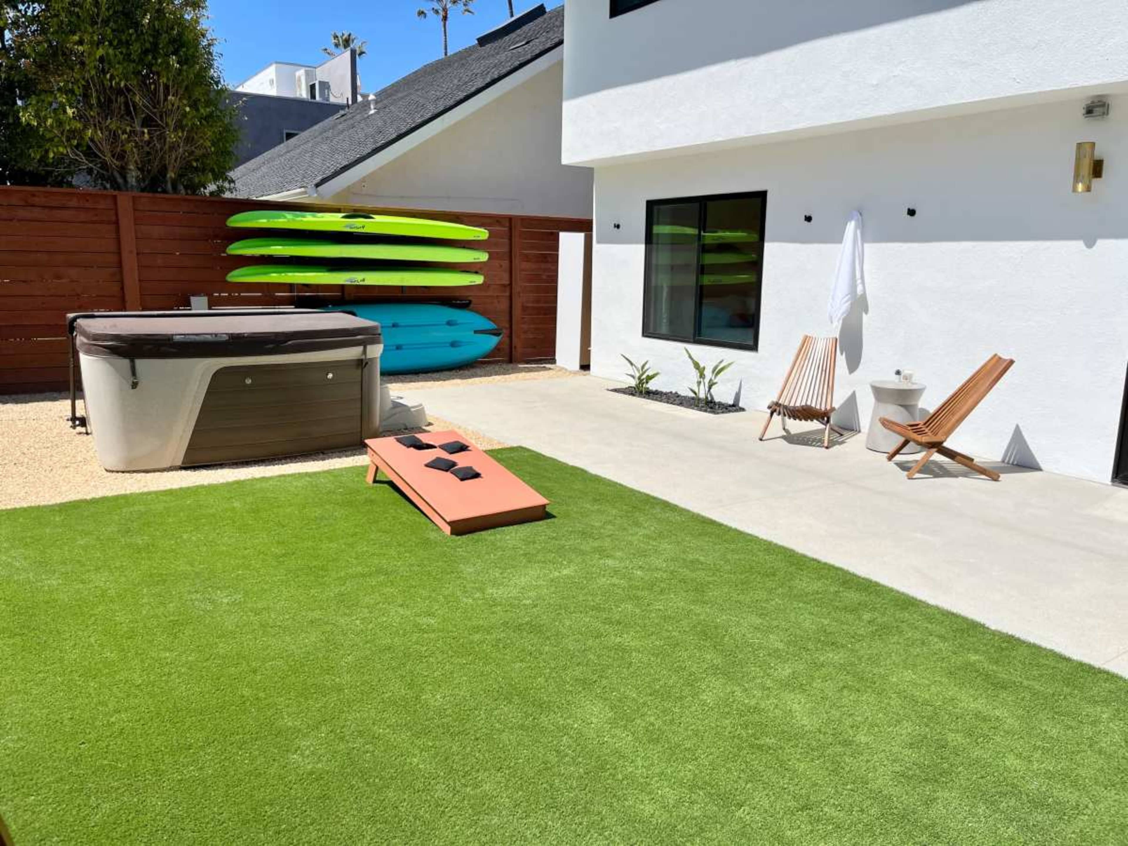 The image features a backyard with a hot tub, two lounge chairs, a small table, and several brightly colored kayaks leaning against a wall.