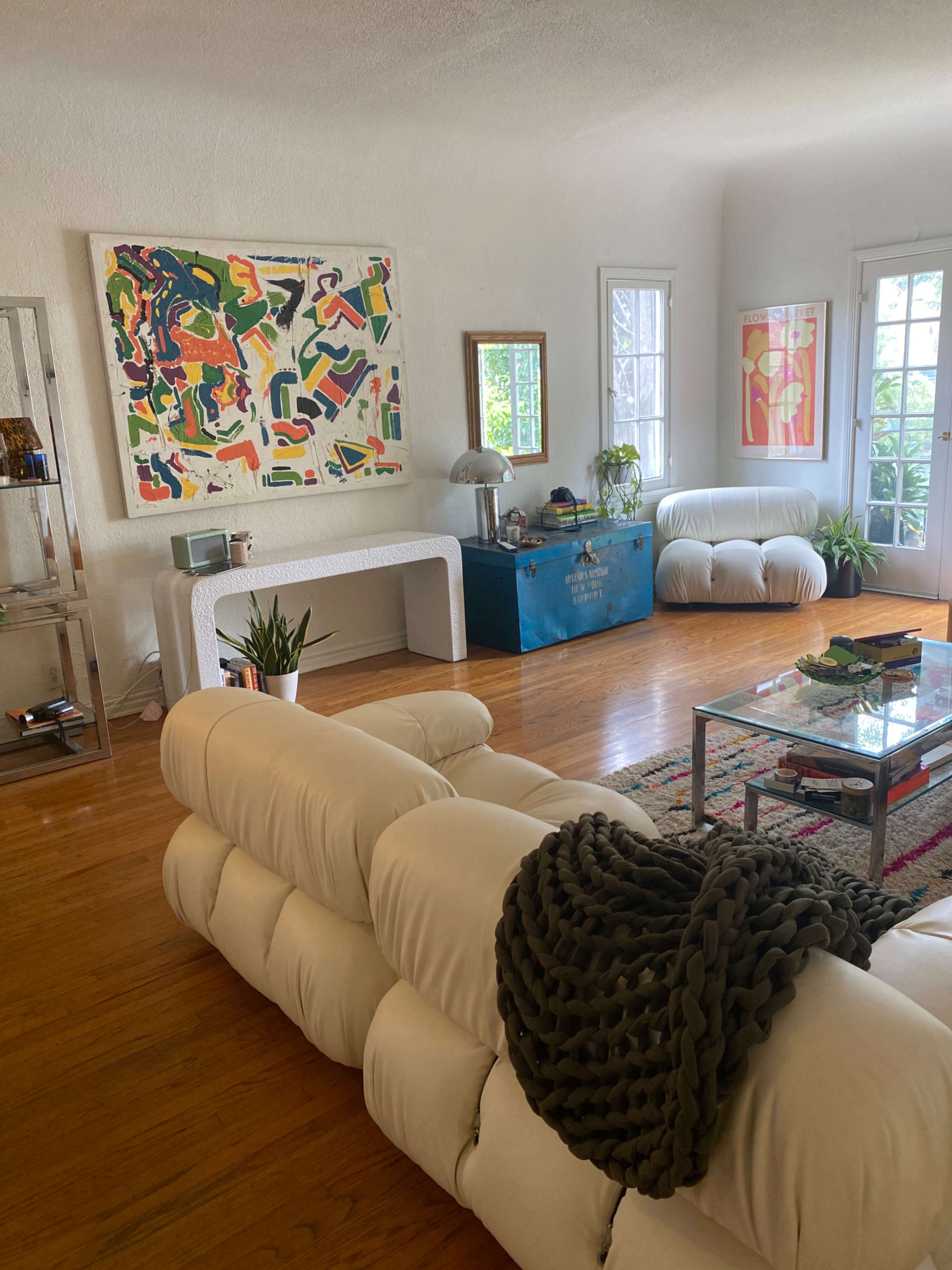 A bright living room featuring a large abstract painting, a white sectional sofa, a light wooden floor, and a blue storage chest against the wall.