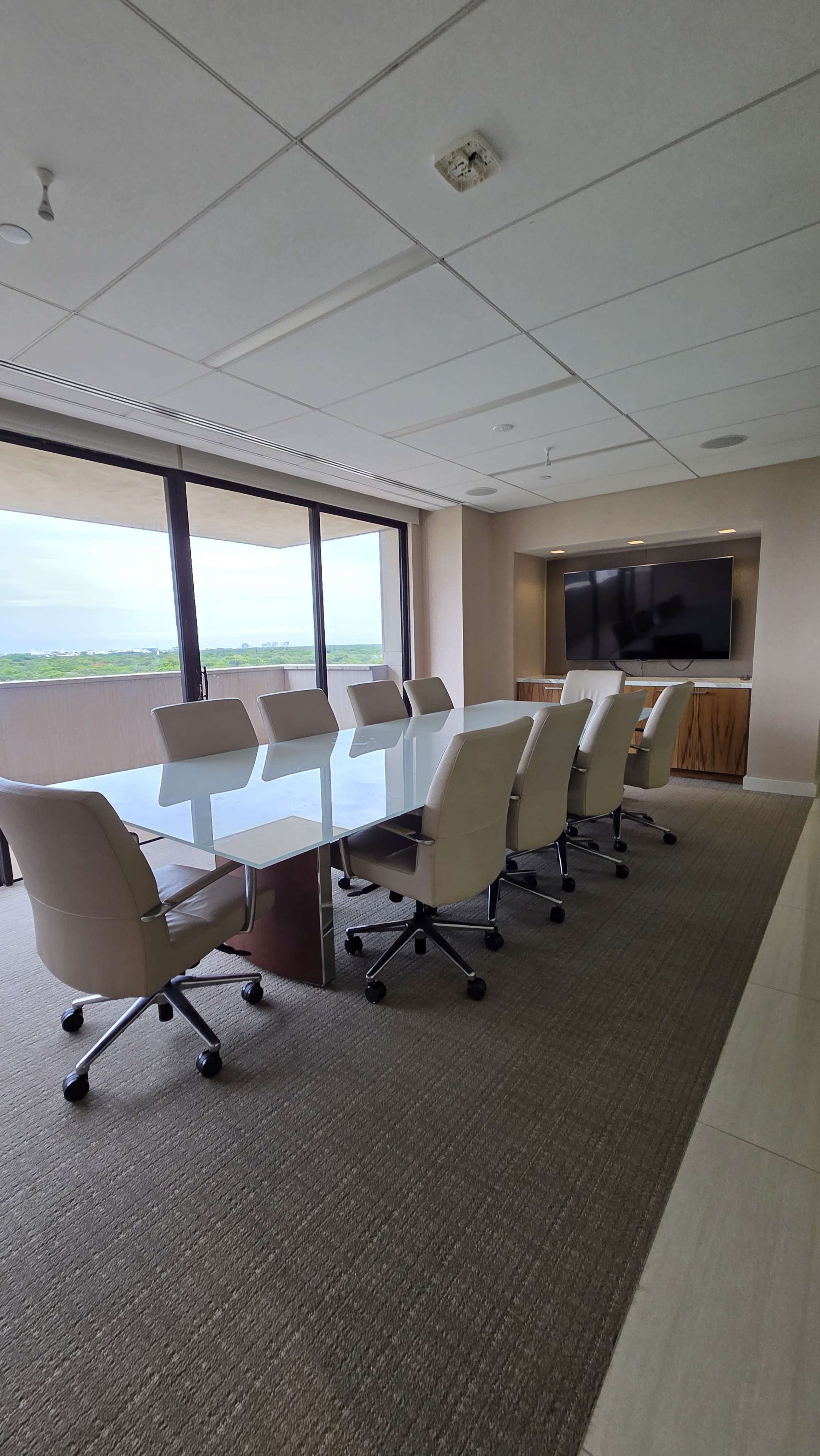 A conference room features a long glass table surrounded by white ergonomic chairs and a large screen mounted on the wall.