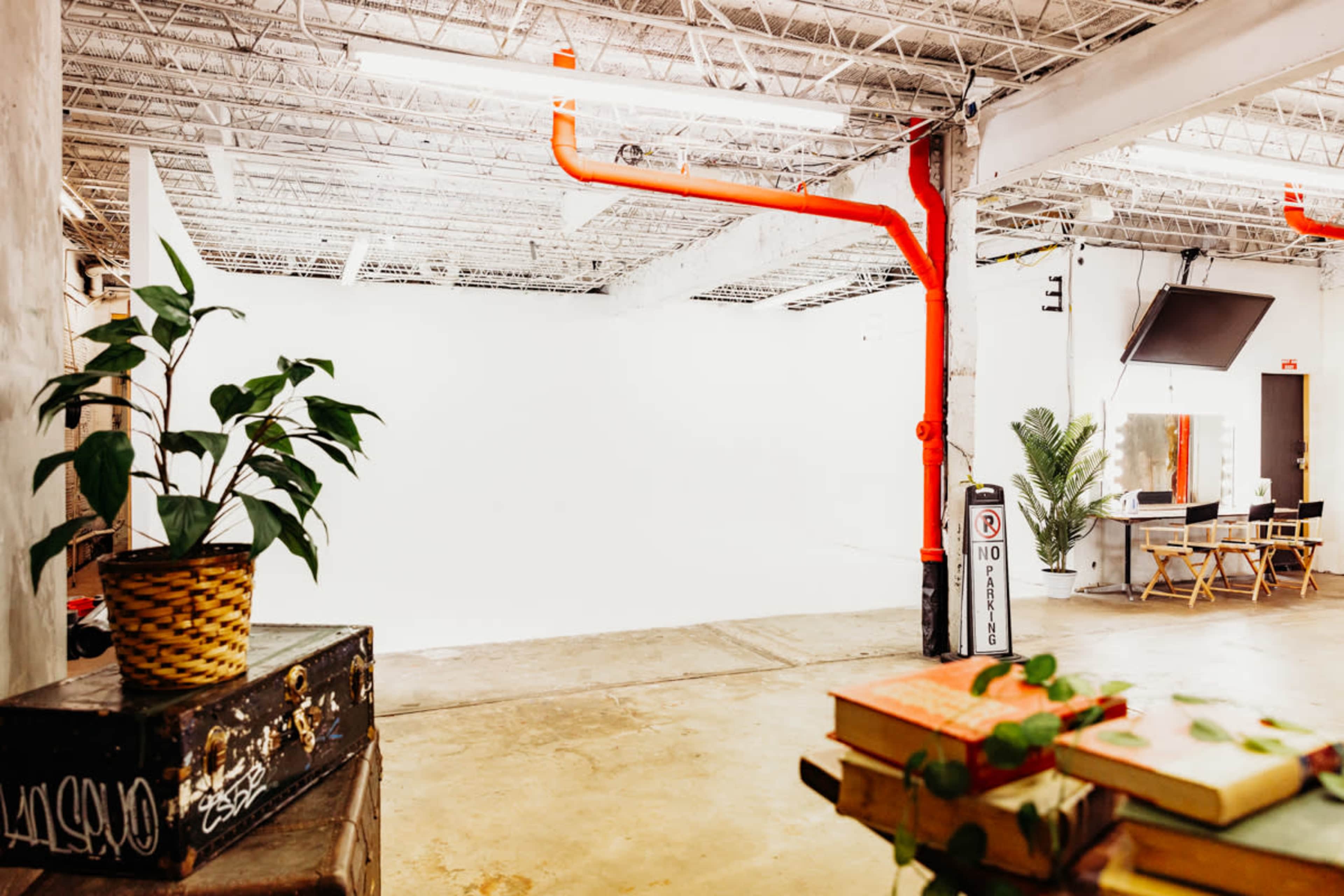 A spacious, well-lit studio with white walls, orange pipes, a plant in a woven basket, and furniture including a table and chairs.