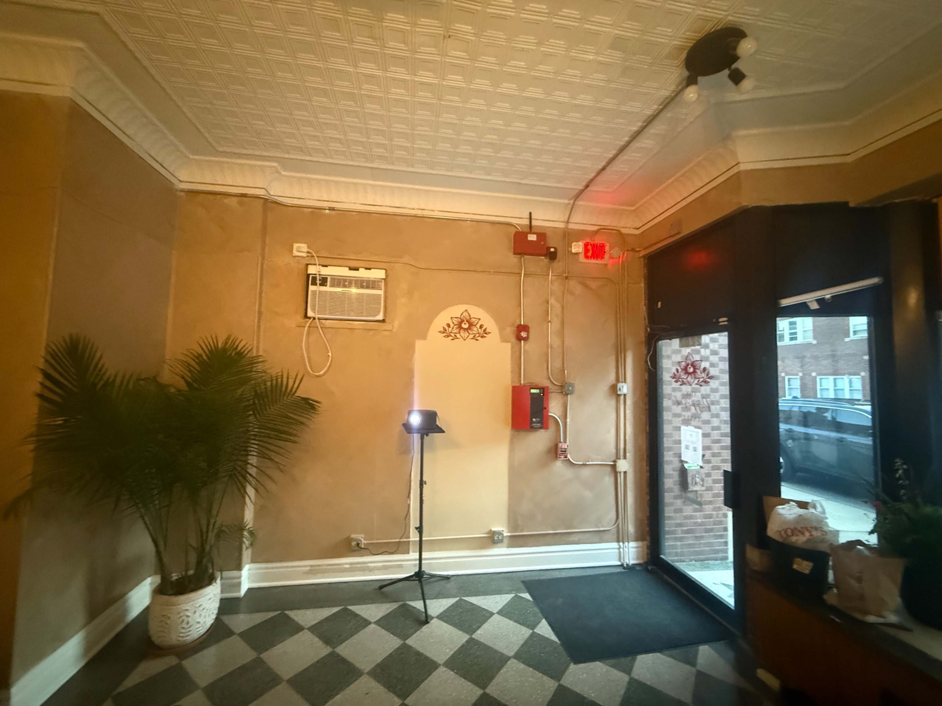 The image shows a lobby area with a potted plant, an air conditioning unit on the wall, and an exit door with red emergency lighting.