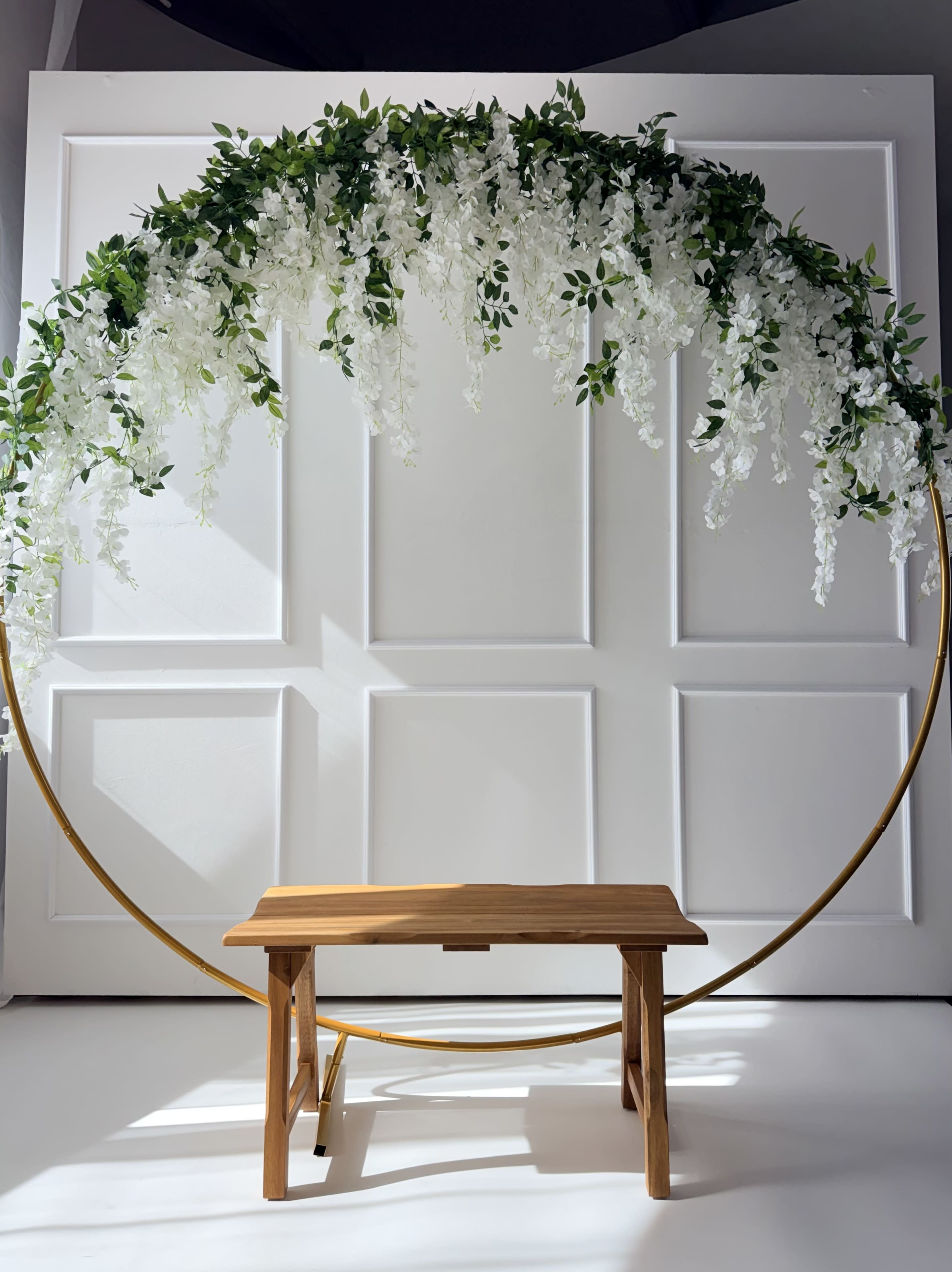 The image shows a circular floral arch adorned with white flowers, positioned above a simple wooden bench against a plain white background.
