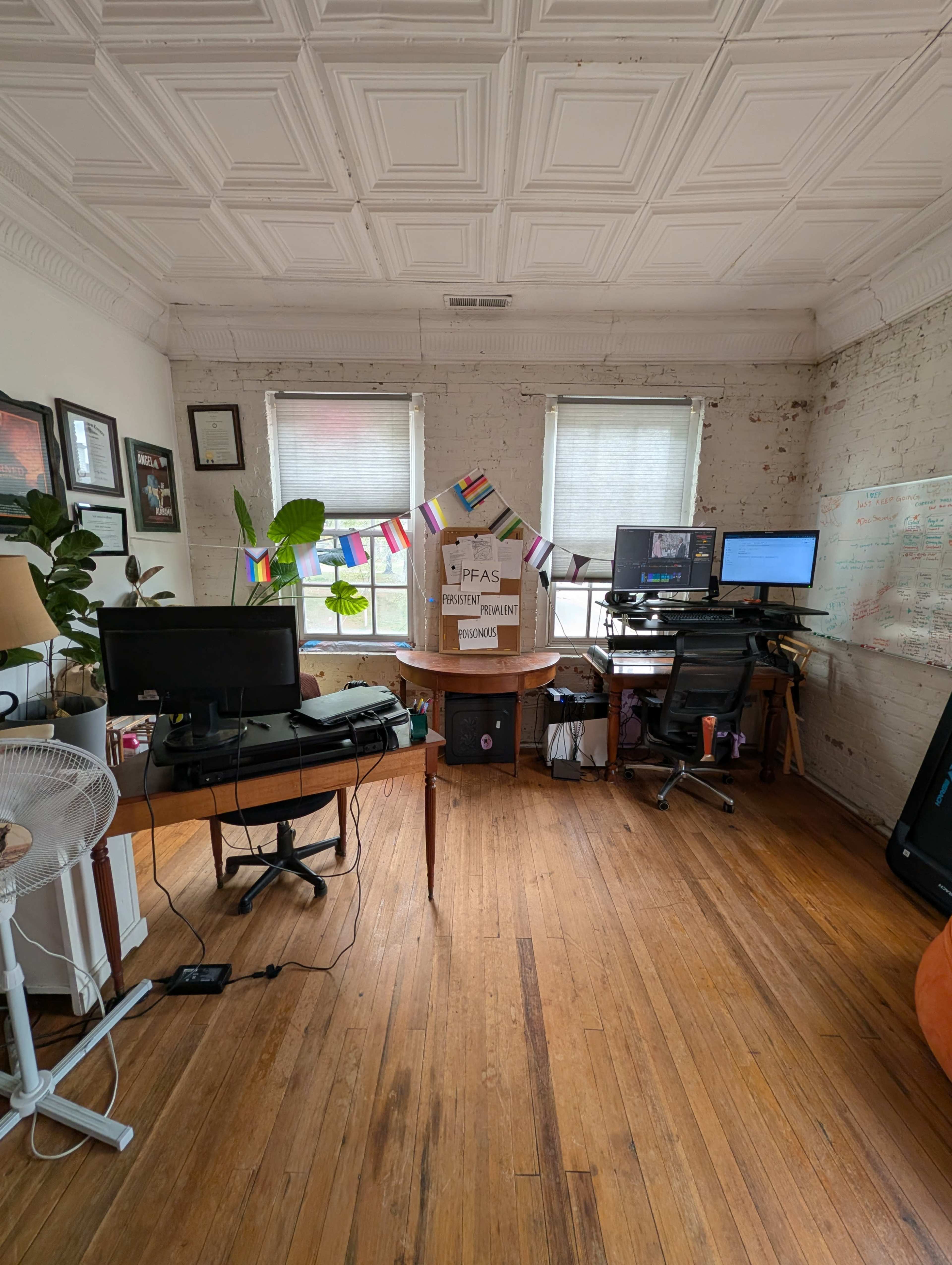 The image shows a workspace with two desks equipped with computers, a central table, and plants, set in a room featuring wood flooring and decorative ceiling tiles.