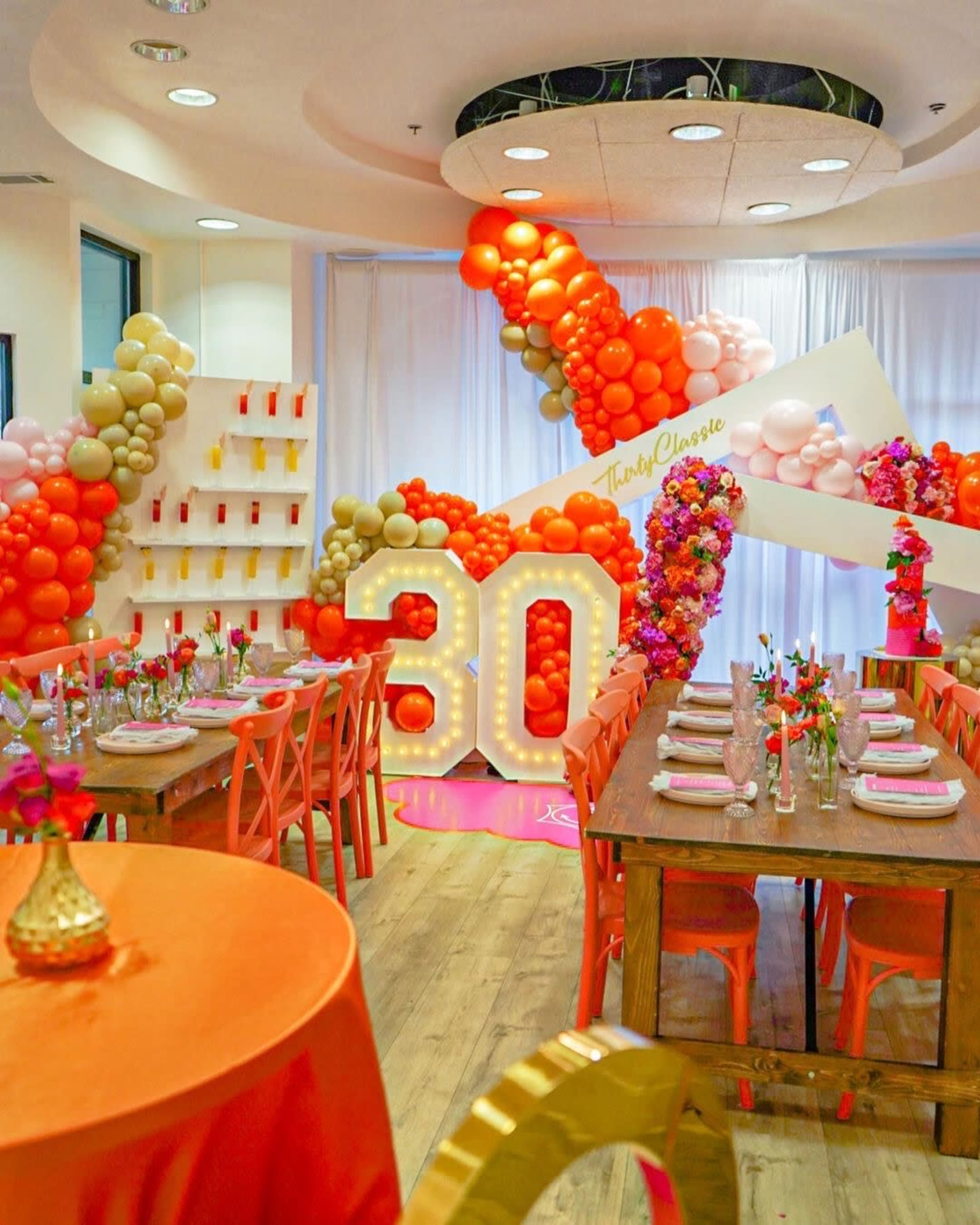 A decorated event space features two large tables with orange tablecloths, surrounded by colorful balloon arrangements and a prominent "30" display.