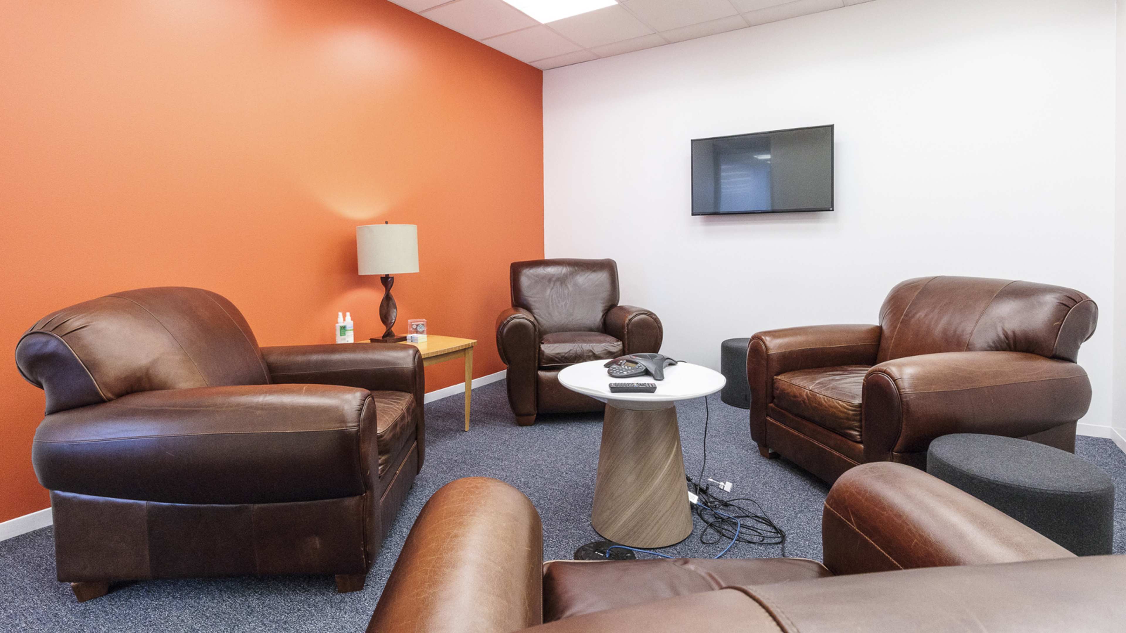The image shows a small lounge area with two leather couches, a coffee table, a lamp, and a wall-mounted television, all set against an orange wall.