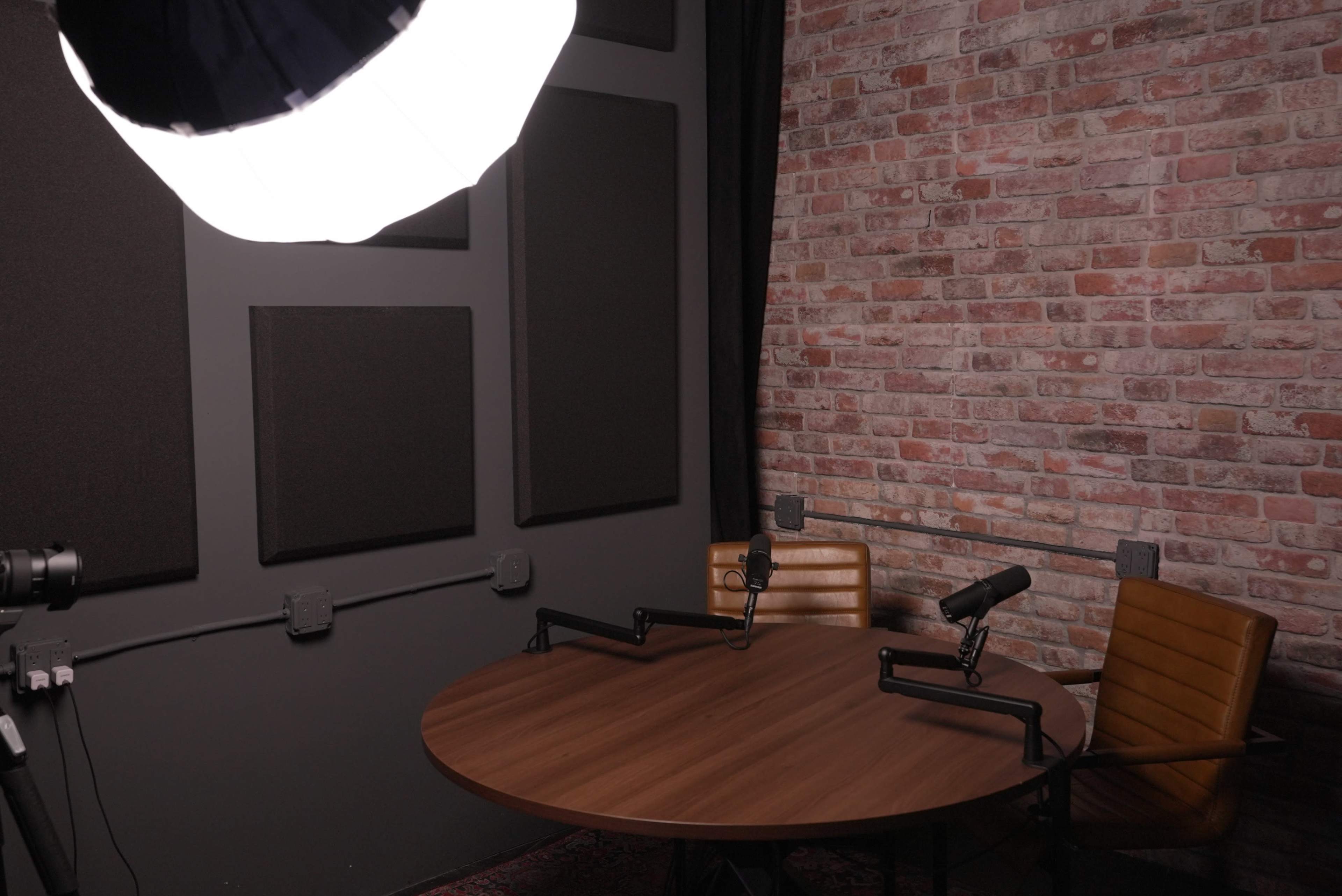 A round wooden table with two chairs and microphones is set up in a room with a brick wall and soft lighting.