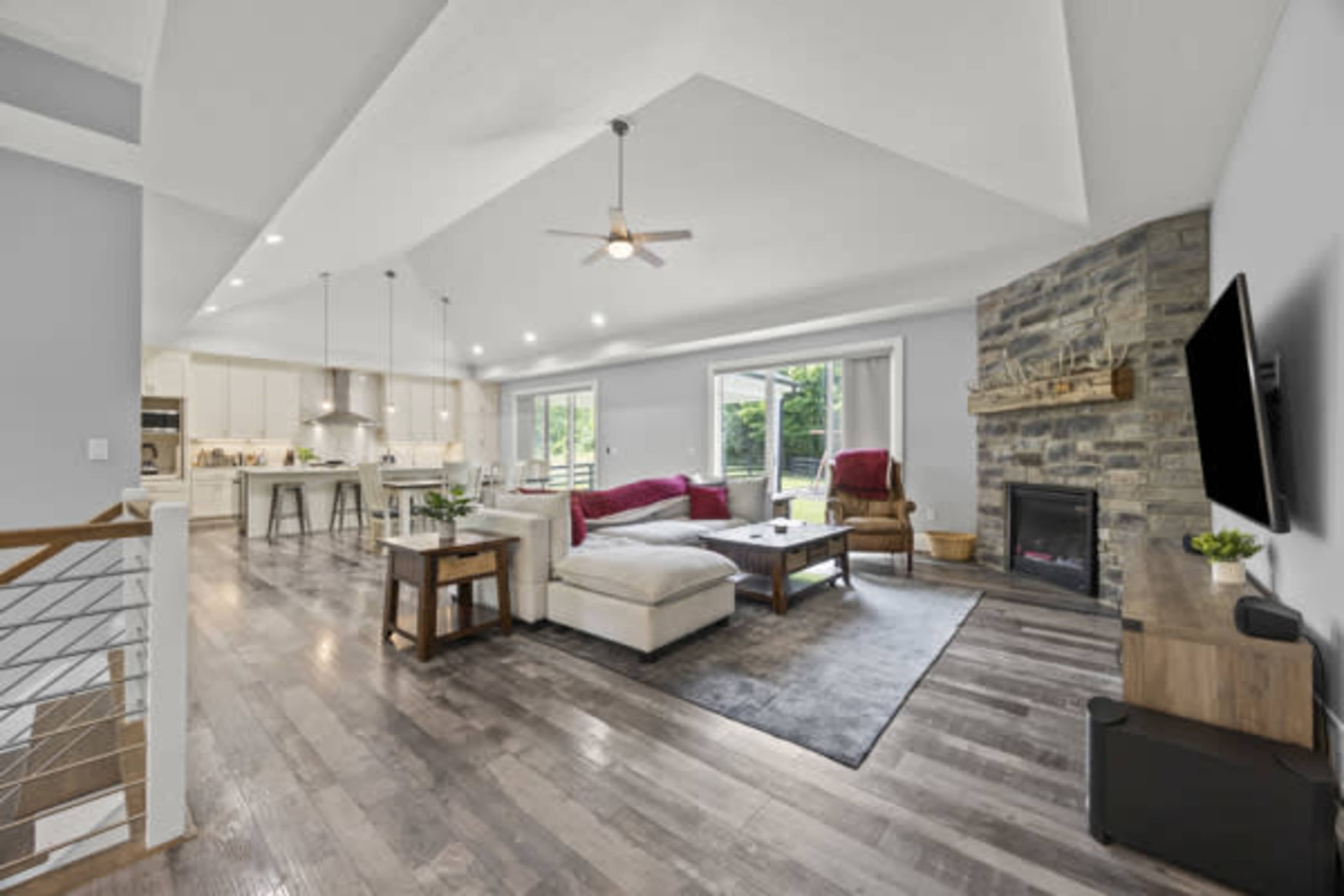 The image shows a modern living room with a stone fireplace, a sectional sofa, and a view of a bright kitchen area in an open layout.