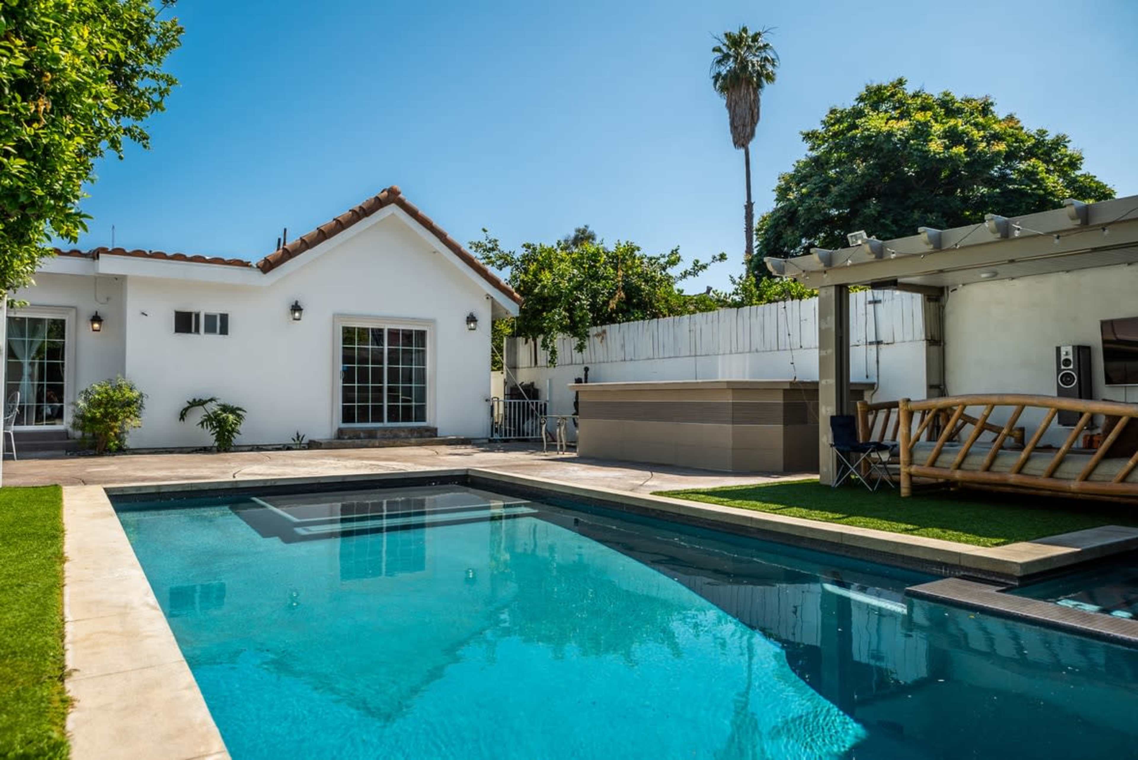A residential backyard featuring a swimming pool, a spa area, a white house with a tiled roof, and landscaped greenery.