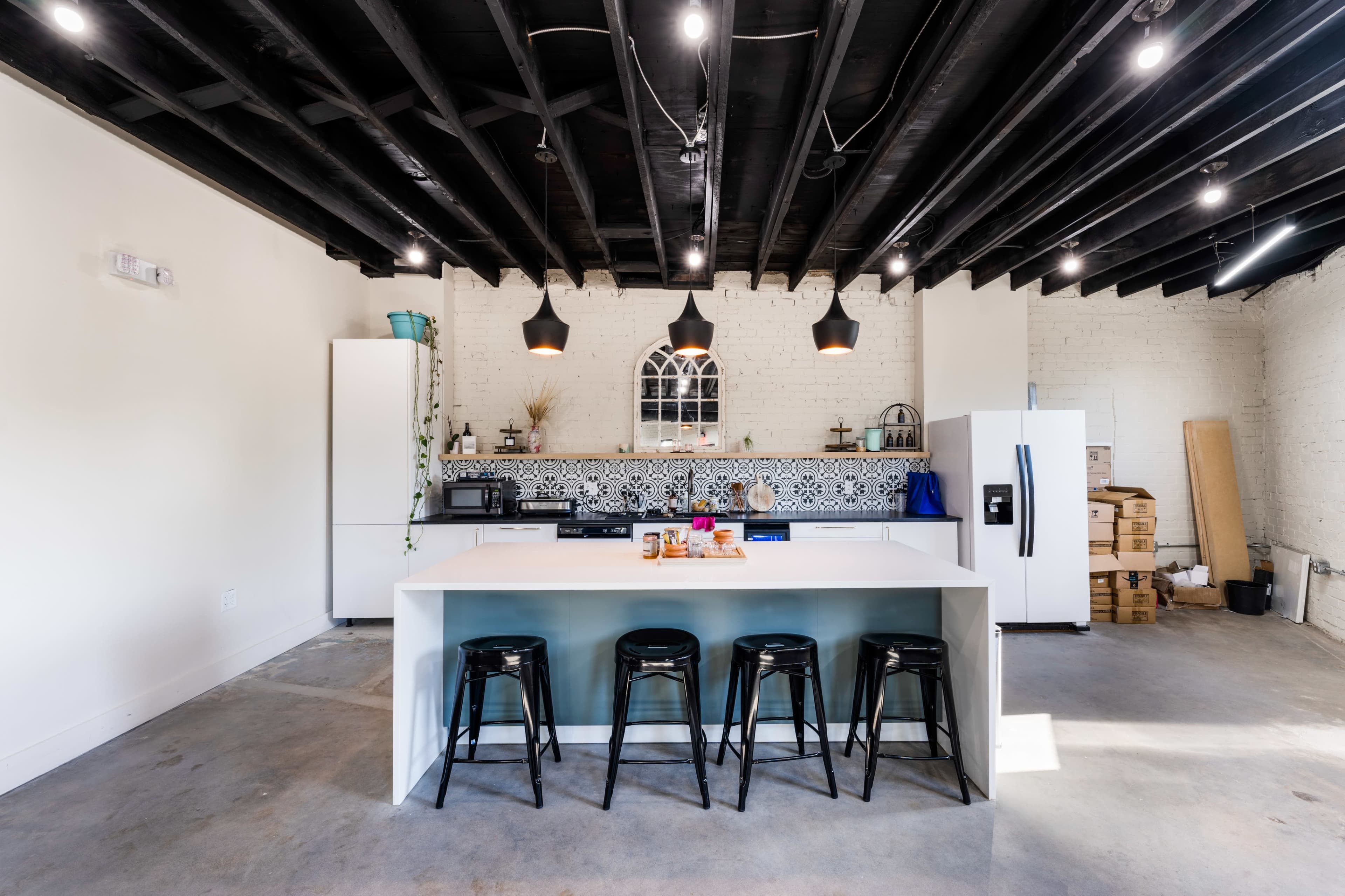 The image shows a modern kitchen with a white island, black bar stools, and a tiled backsplash, illuminated by pendant lights.