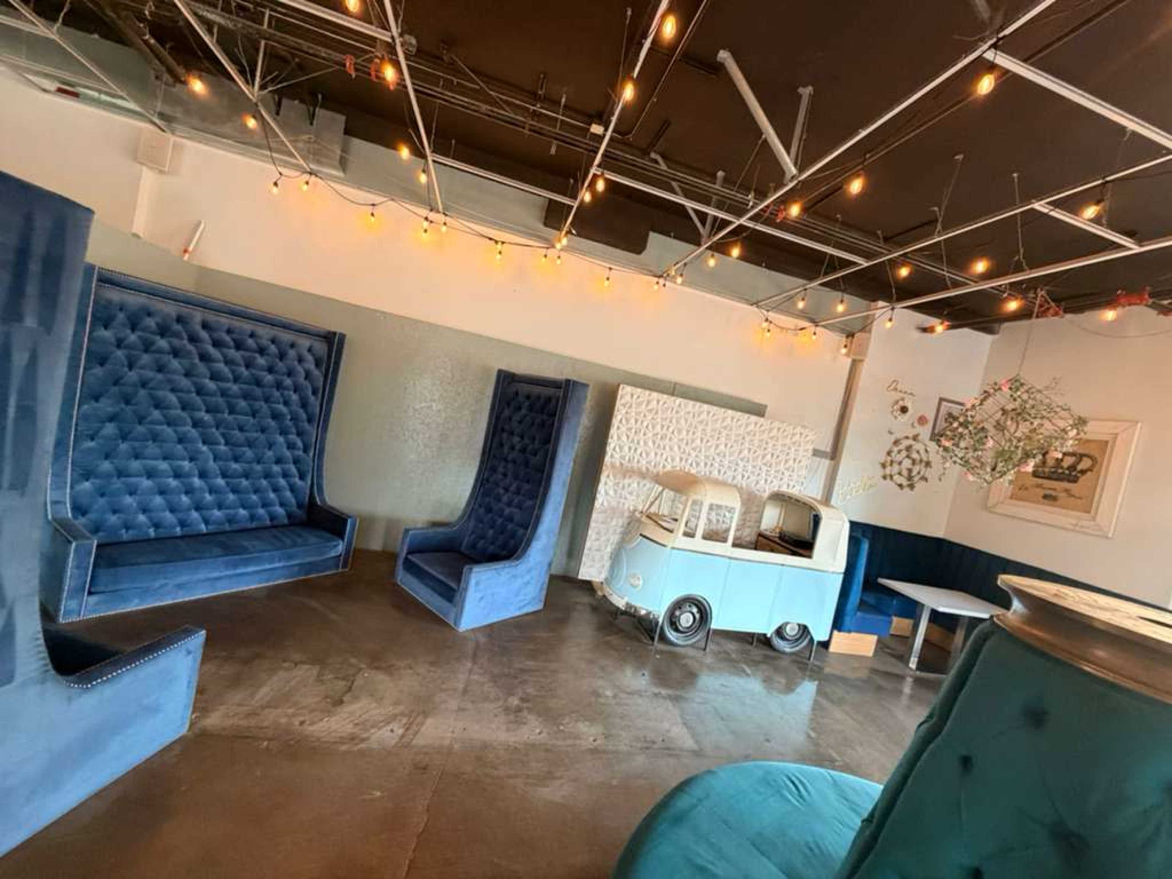 The image shows a stylish interior of a lounge featuring blue velvet seating, ambient string lights, and a decorative vintage car.