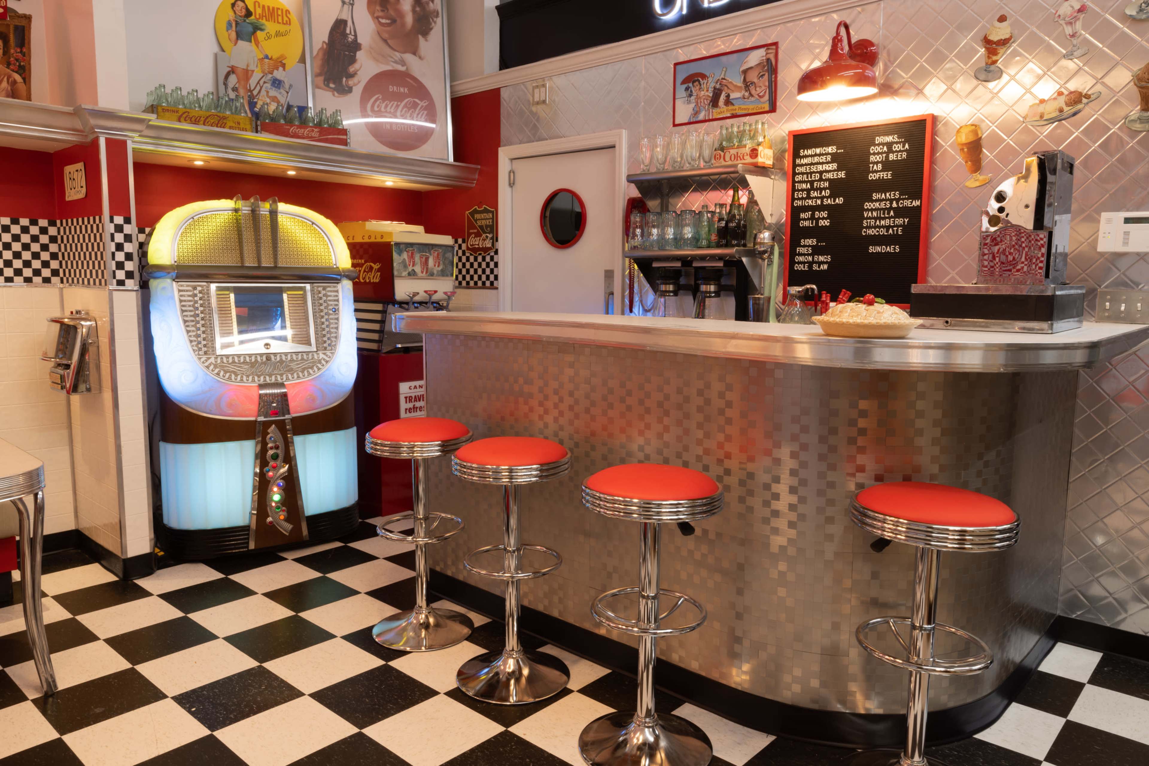 A retro-style diner with a jukebox, a metal counter with red stools, and a black and white checkered floor.