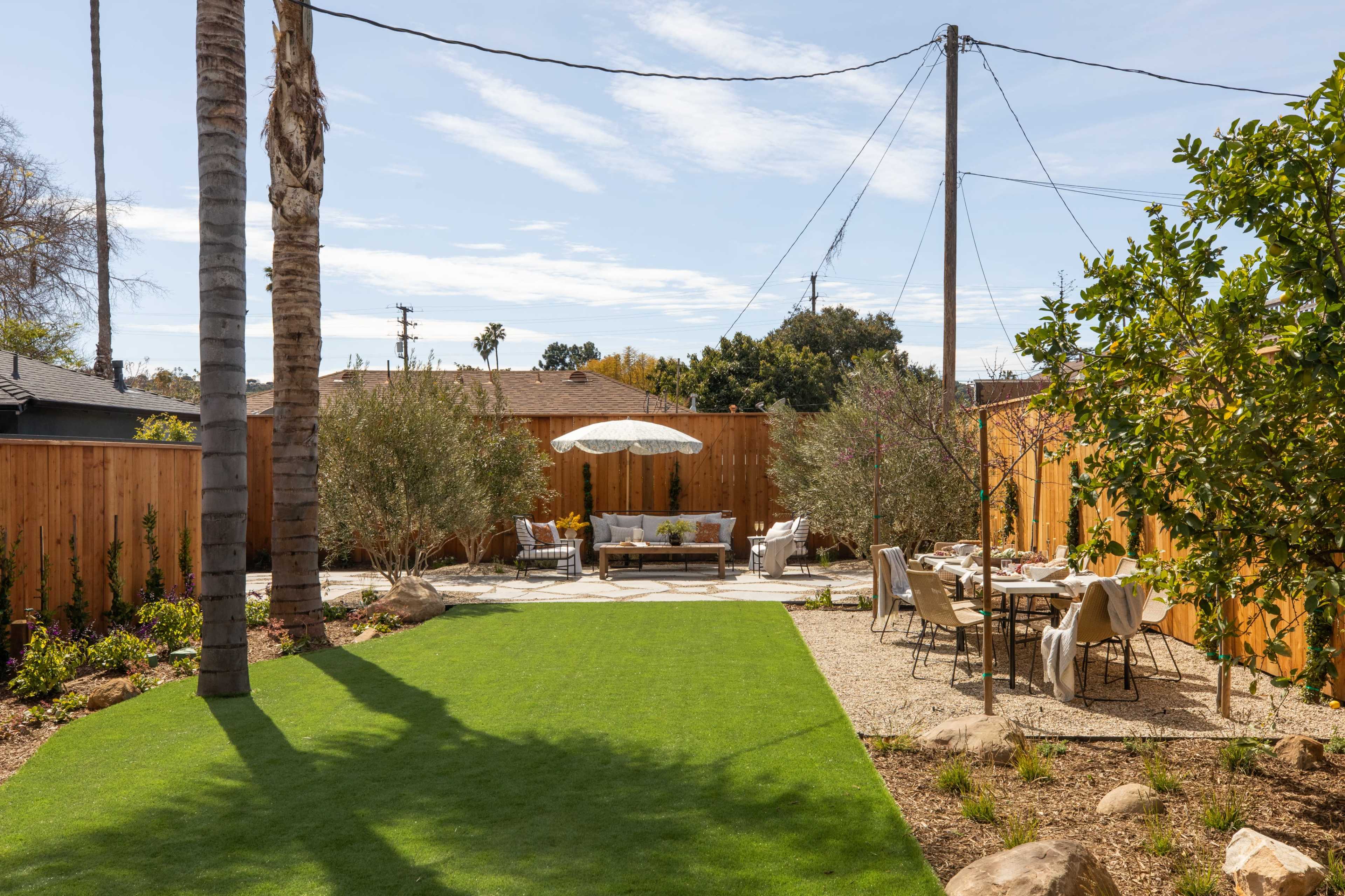A landscaped backyard featuring a green lawn, a dining area with a table and chairs, and a seating area shaded by an umbrella, all surrounded by wooden fencing and decorative plants.