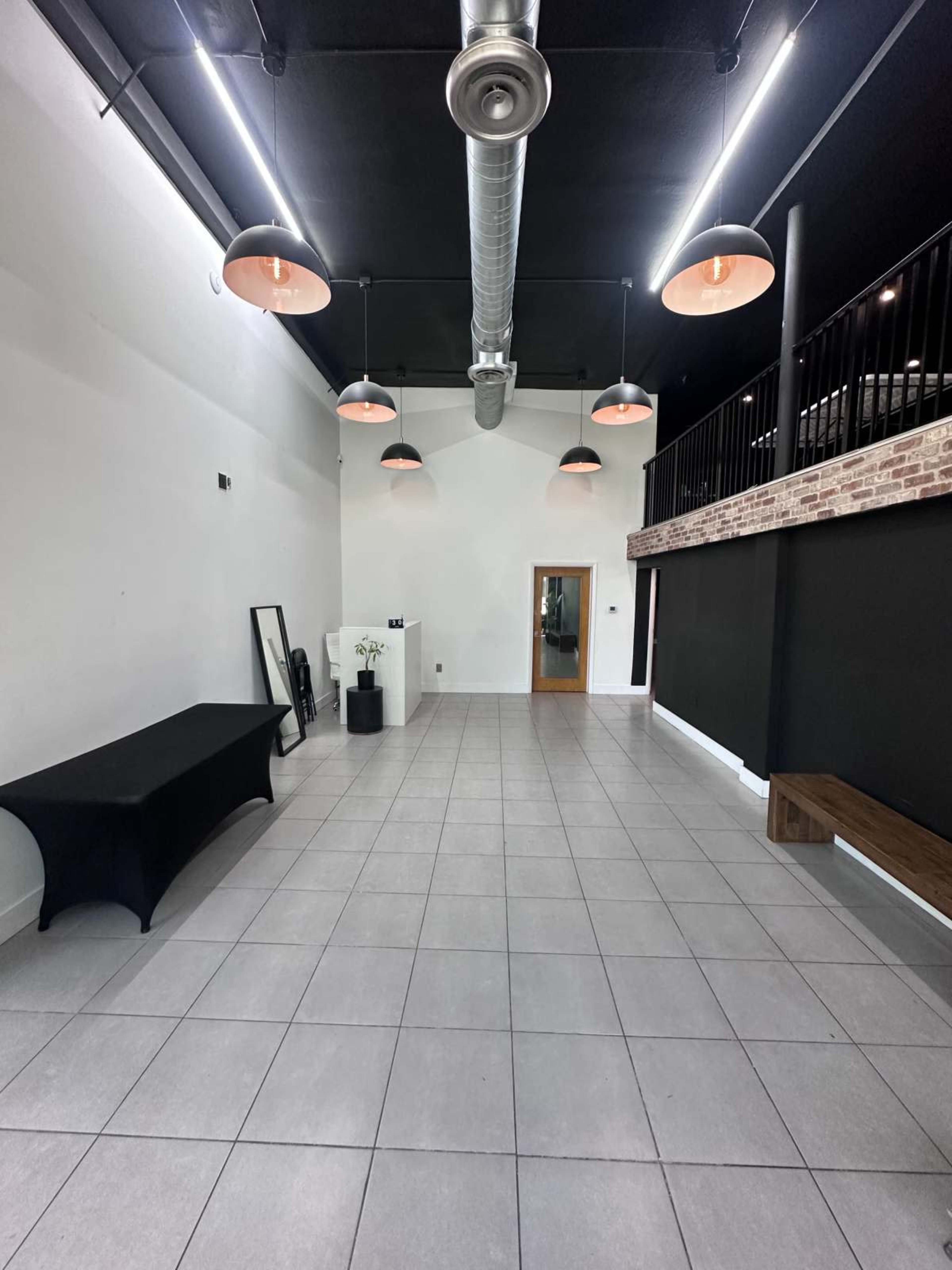 The image shows a spacious, well-lit interior with high ceilings, featuring a black table, a white wall, and a balcony area.
