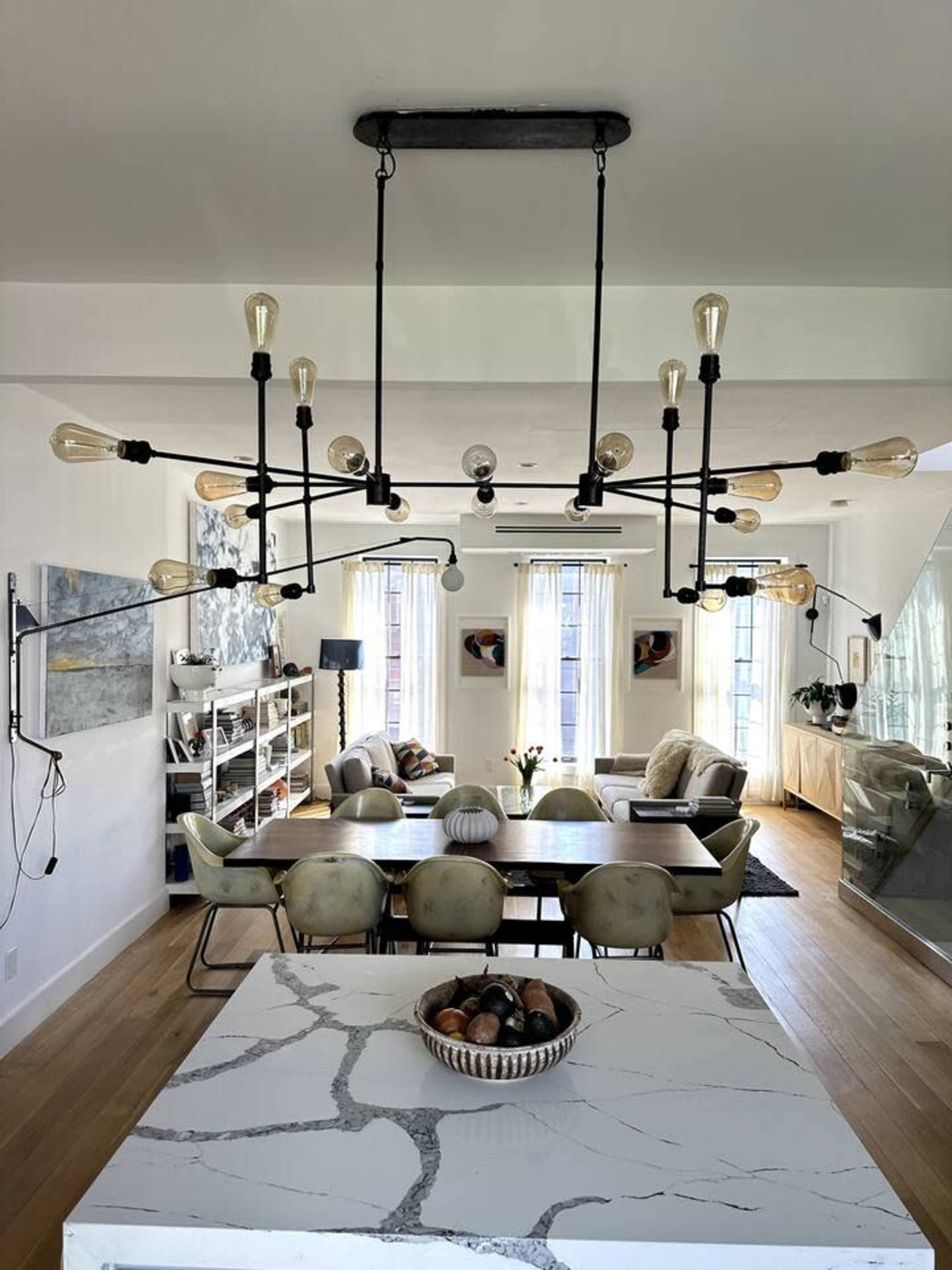 A modern dining area with a large table, surrounded by chairs, and an industrial-style chandelier hanging above, with a kitchen island featuring a marble top in the foreground.