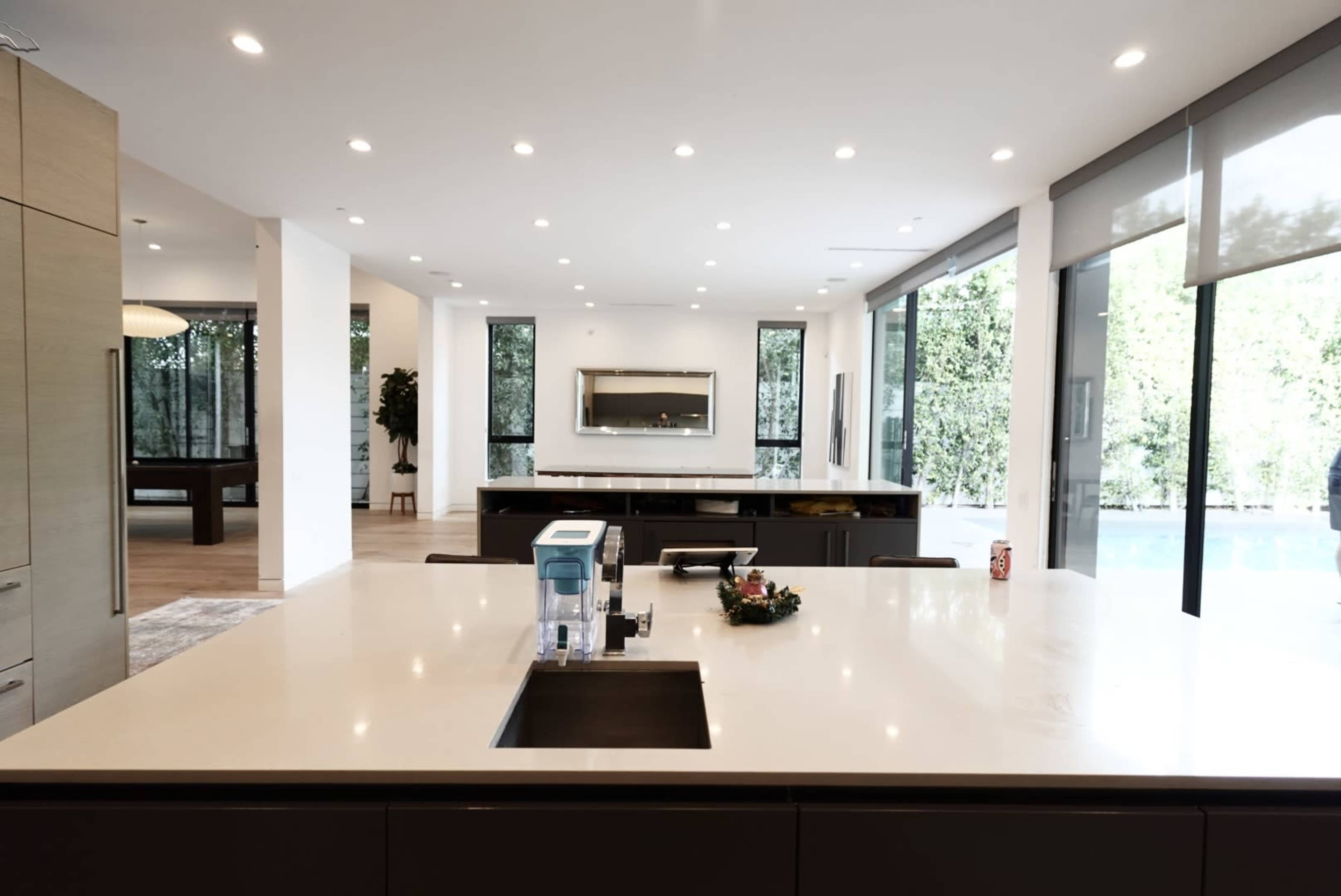 A spacious modern kitchen features sleek cabinetry, a central island with a sink, and large windows overlooking a patio and greenery.