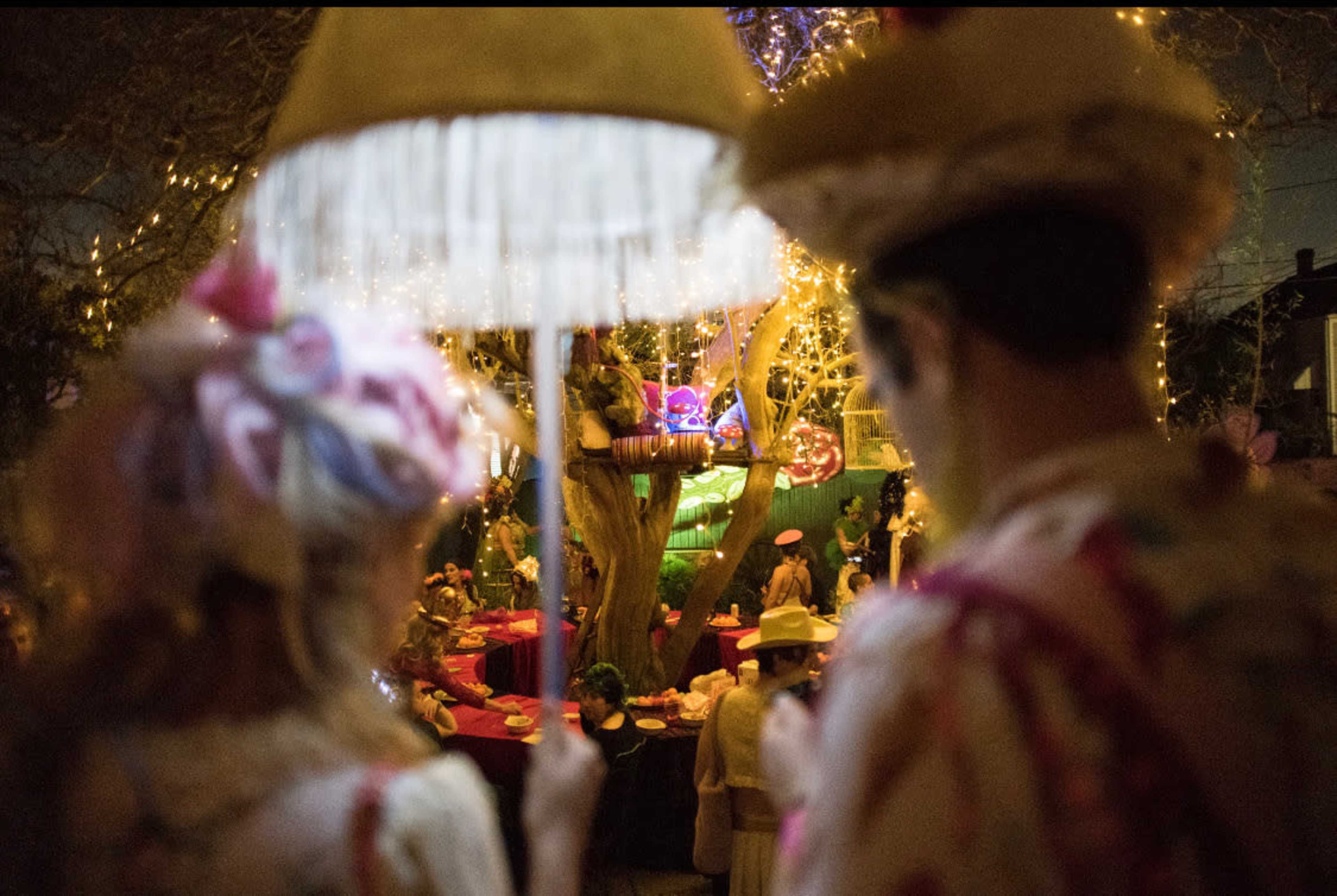 The image shows a festive outdoor gathering with people in colorful attire, illuminated by string lights and decorations around a large tree.