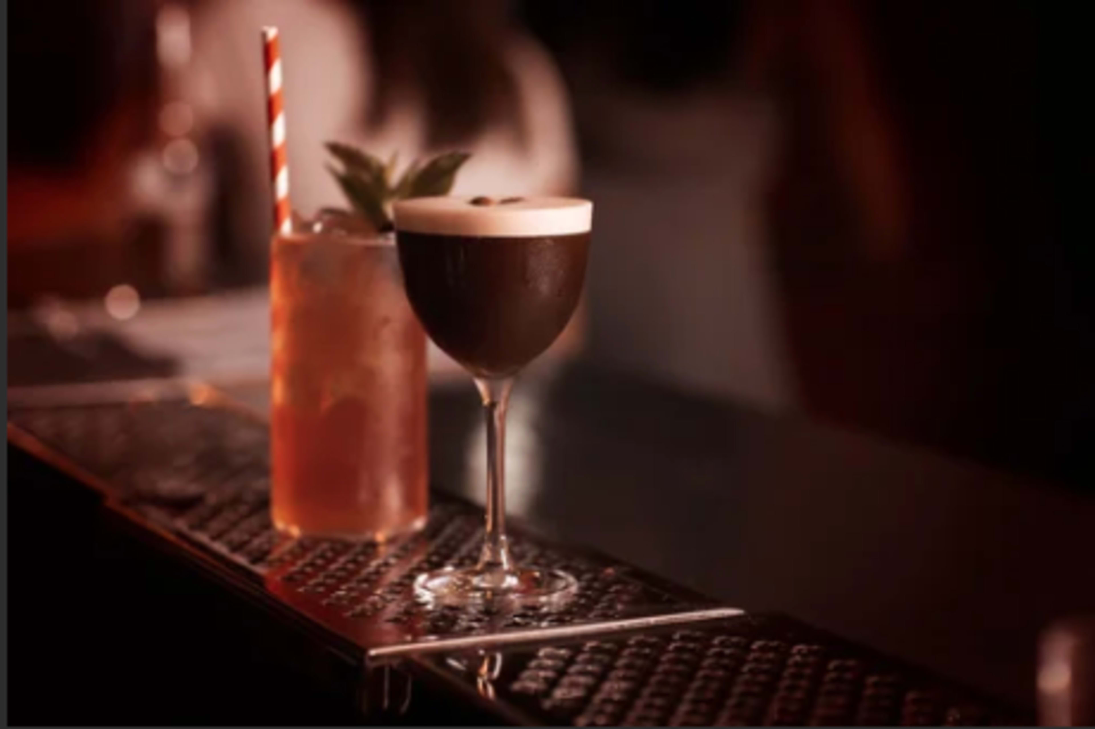 The image shows two cocktails on a bar counter, one in a tall glass with a striped straw and the other in a stemmed glass with a frothy top.