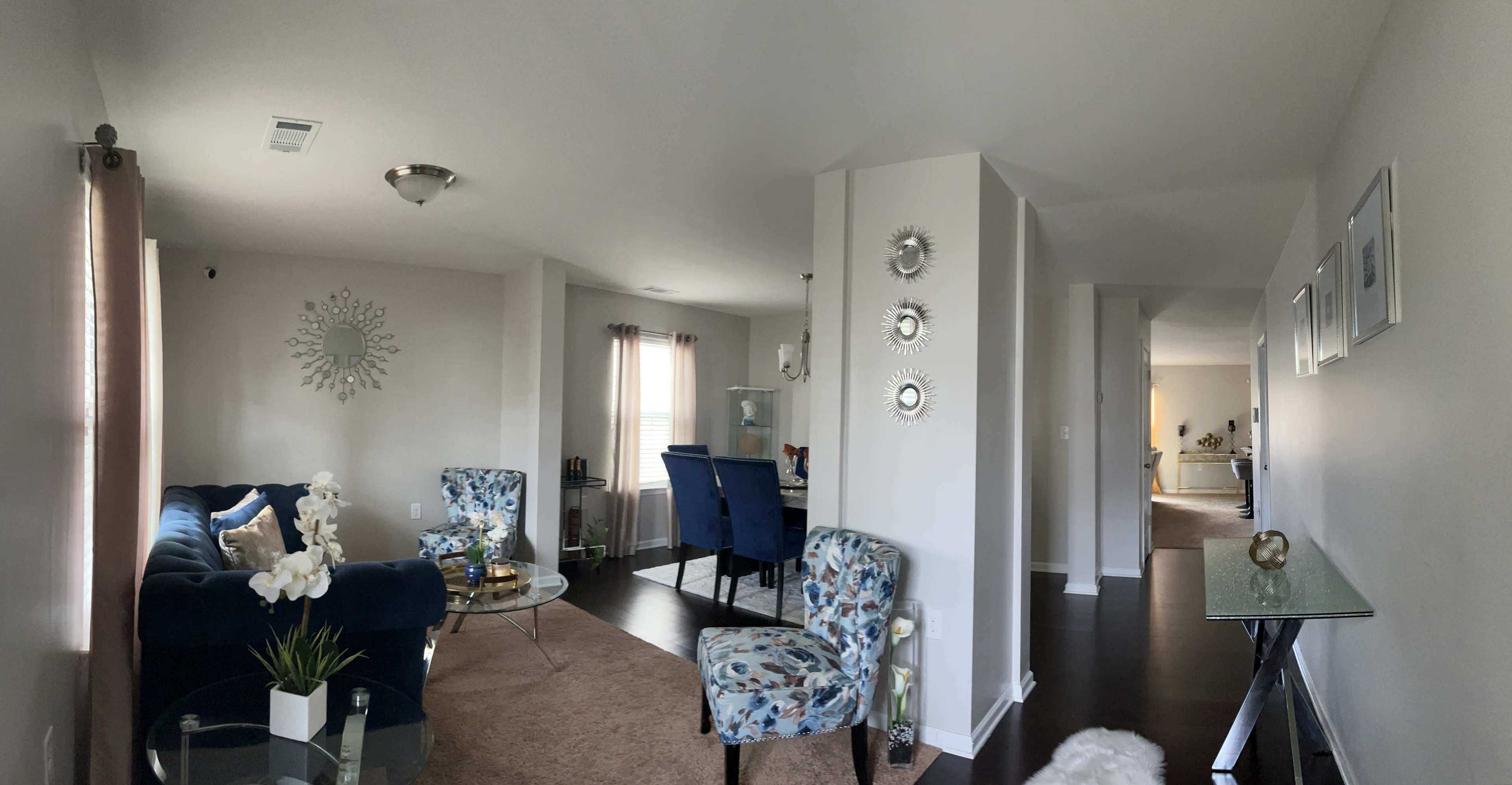 The image shows a modern living room with blue chairs and a glass table, leading into a well-lit hallway with additional chairs visible in the background.