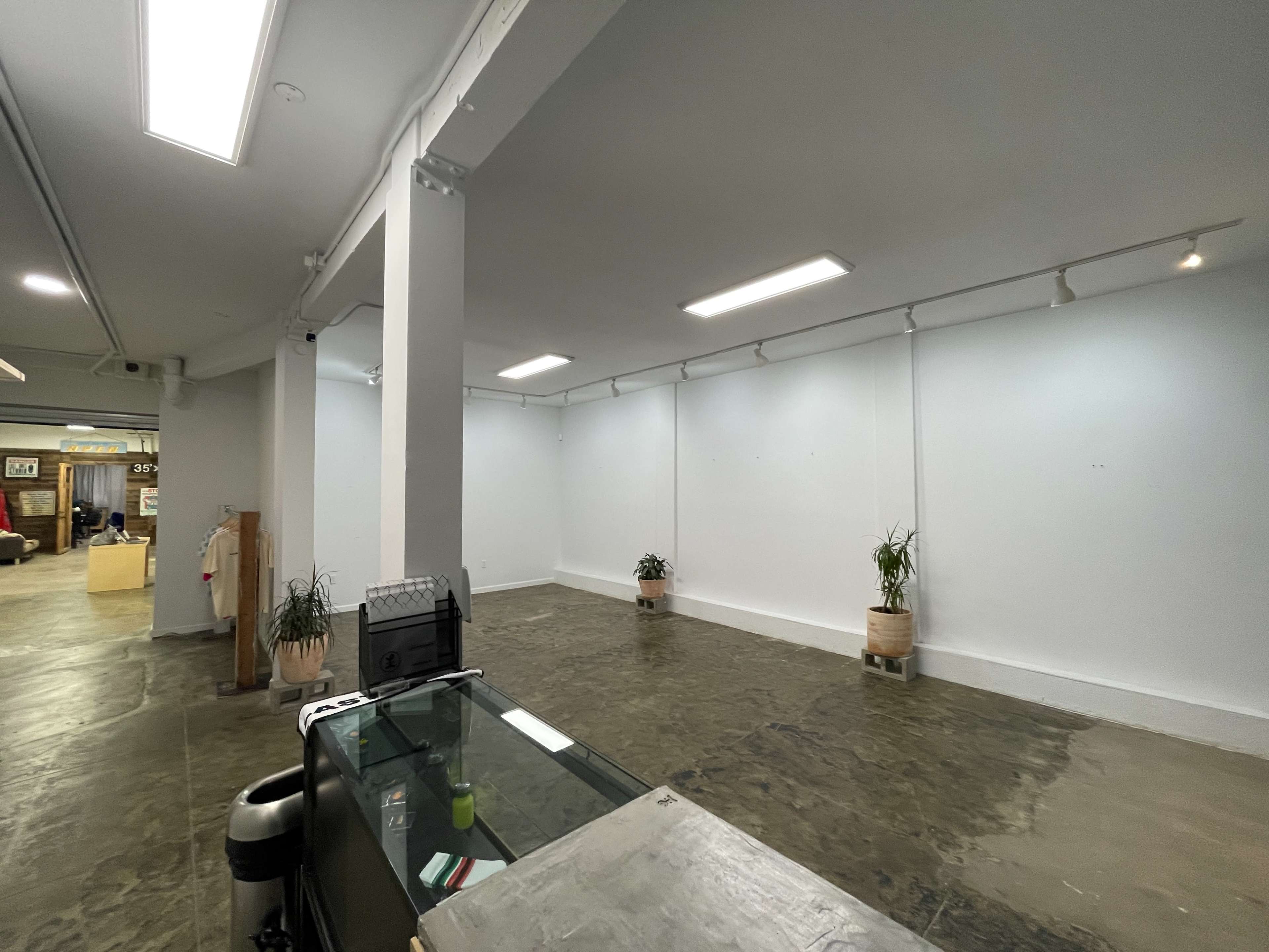 An empty indoor space with concrete flooring, white walls, and a reception area featuring a glass counter and potted plants.