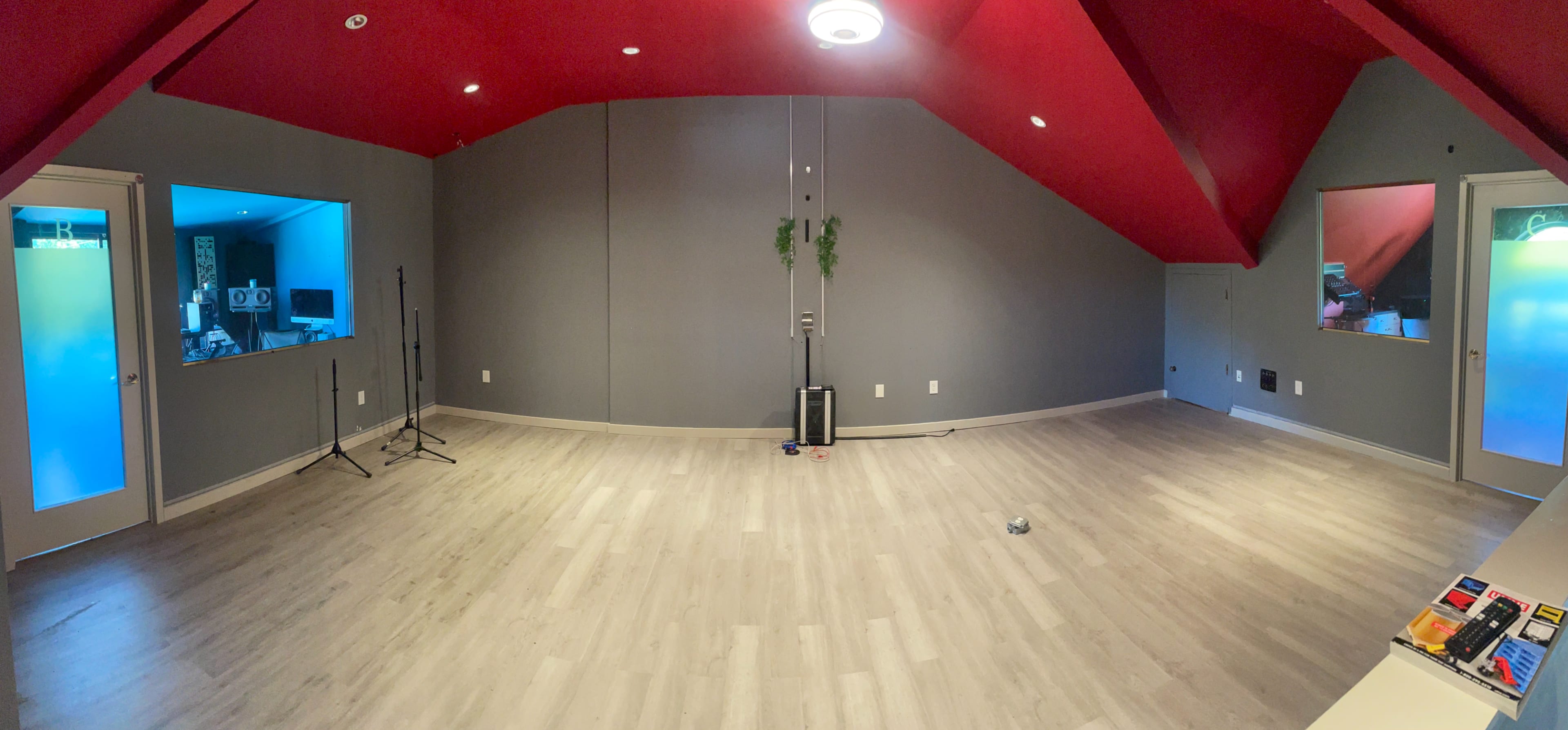 A spacious room with grey walls, a red ceiling, and two small windows, featuring light-colored wooden flooring and a few audio equipment stands.