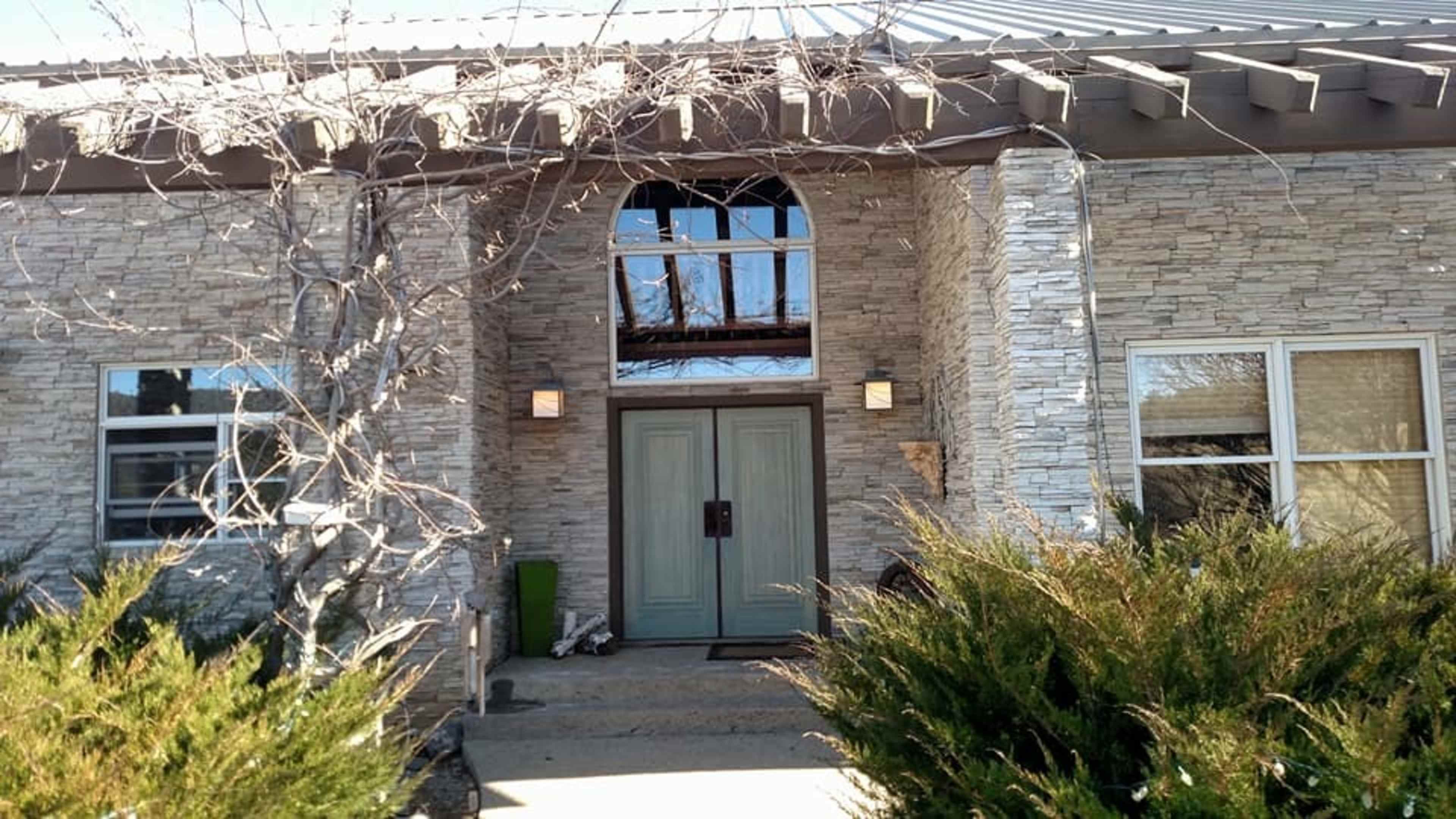 The entrance of a stone-faced house with a pair of green doors and large windows flanking the doorway.