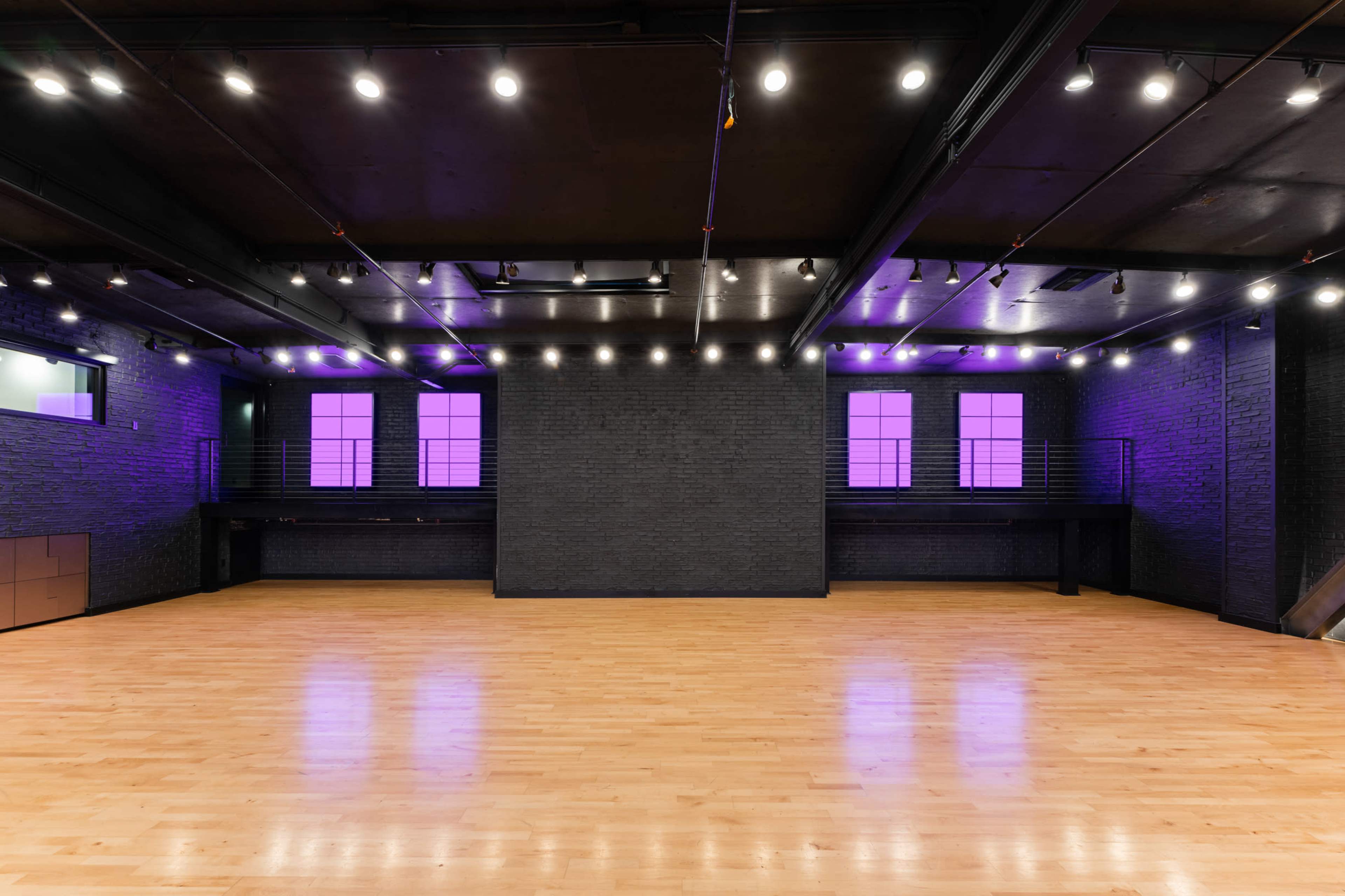A spacious dance studio features a wooden floor and purple-lit walls with large windows, under bright overhead lighting.