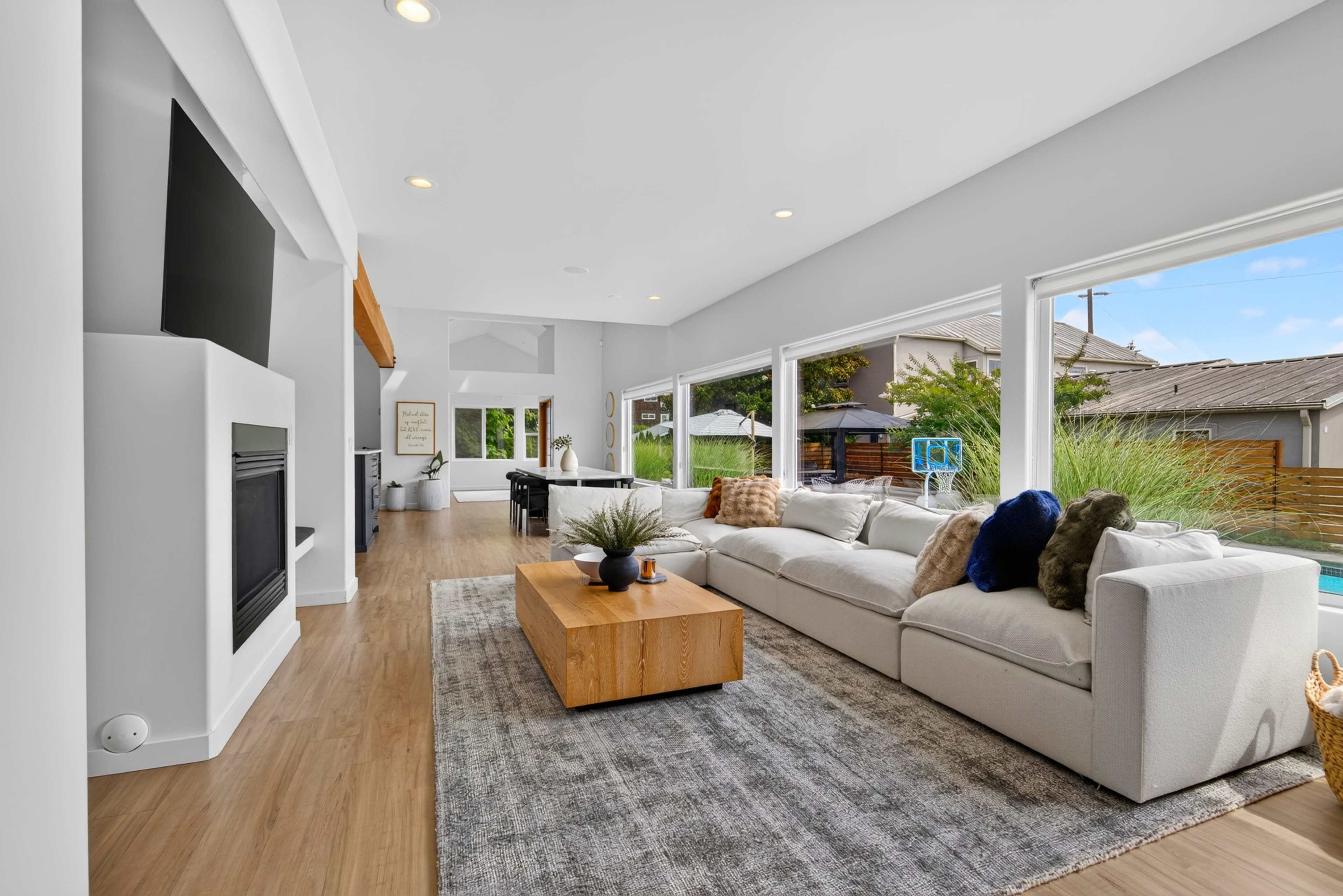 The image shows a modern living room featuring a large sectional sofa, a wooden coffee table, and large windows overlooking an outdoor area.