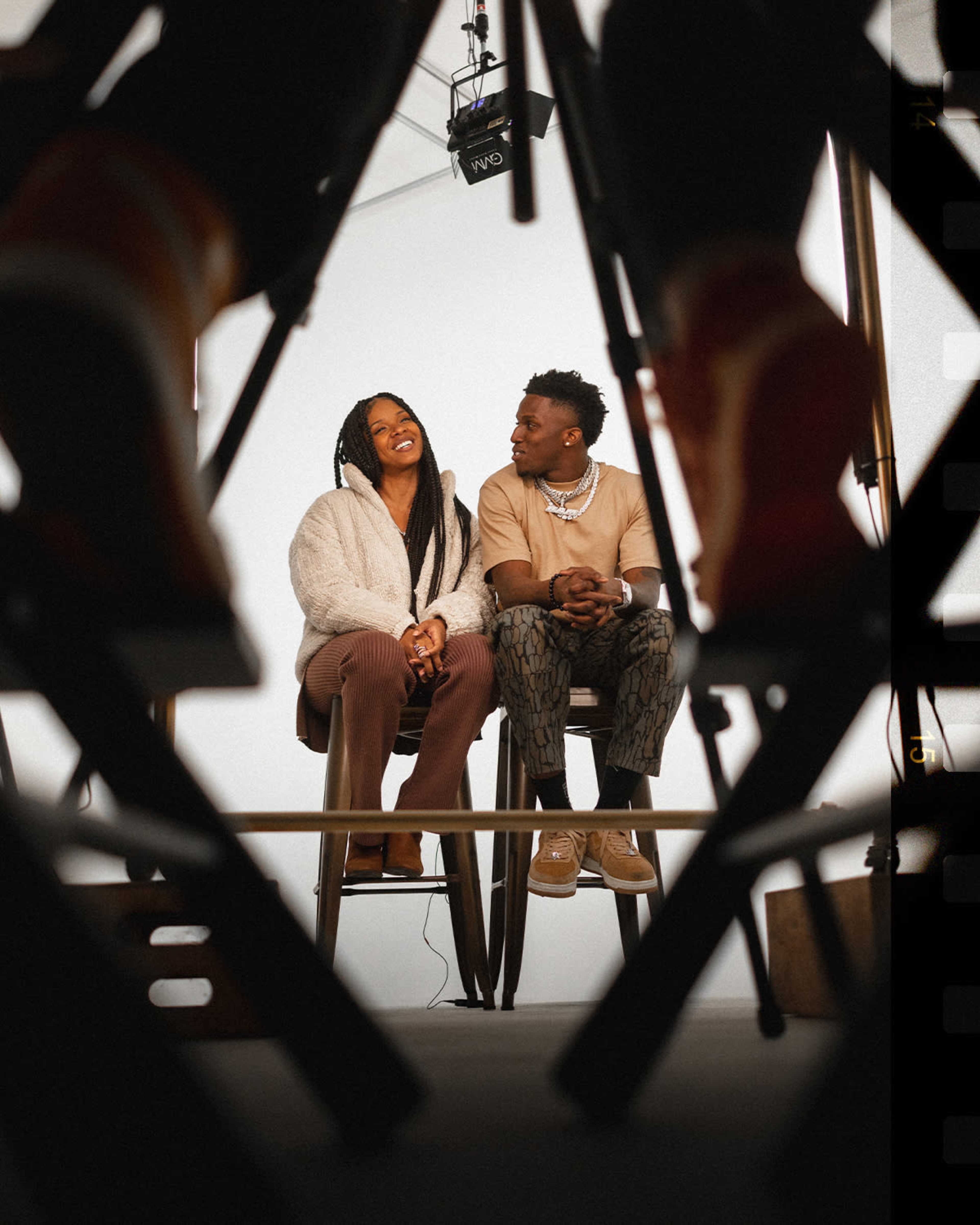 A man and a woman sit on high stools, engaged in conversation, framed by the legs of chairs in the foreground.