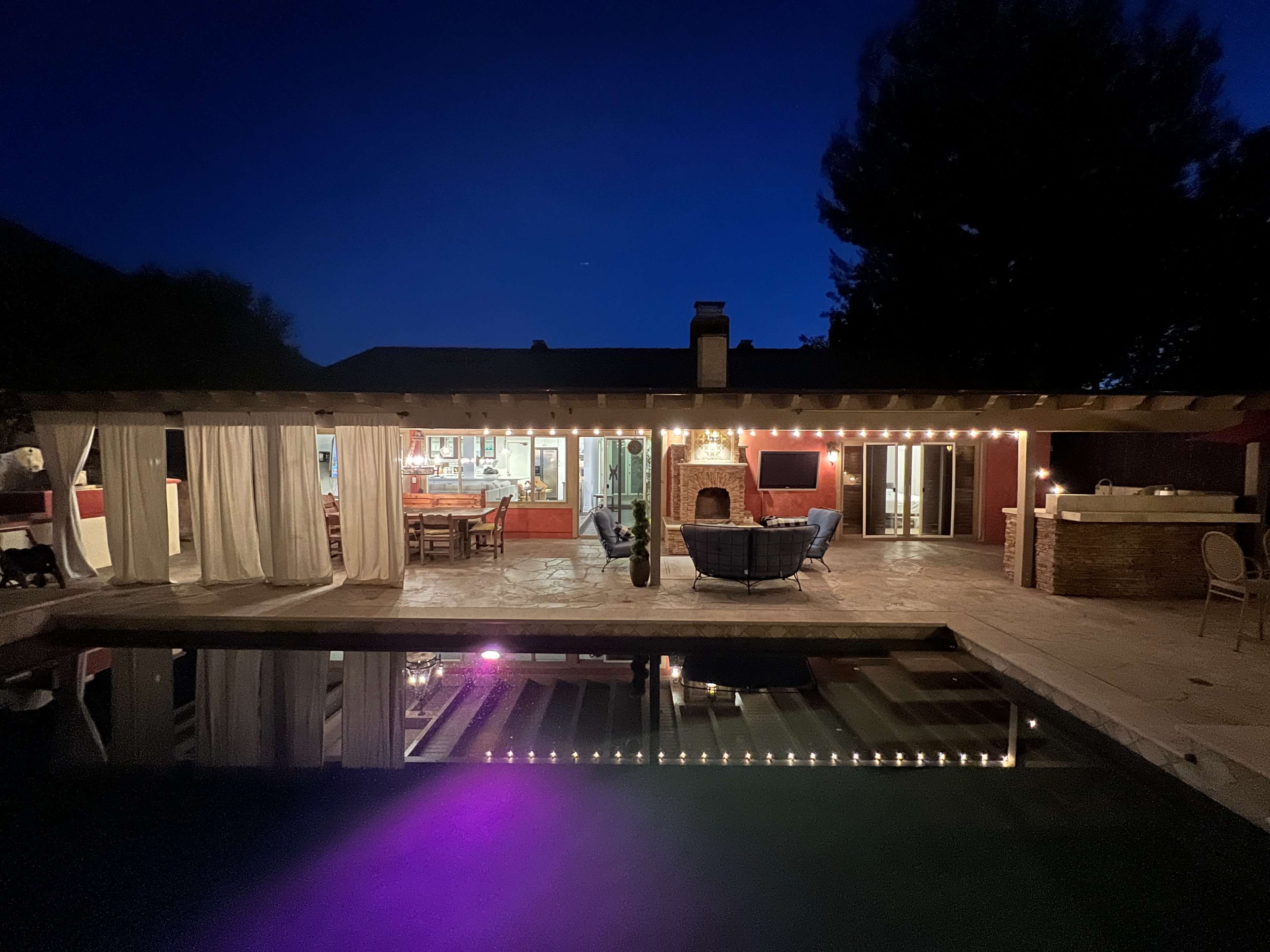 A pool area features a covered patio with string lights and a cozy seating arrangement, reflecting softly in the water beneath a night sky.
