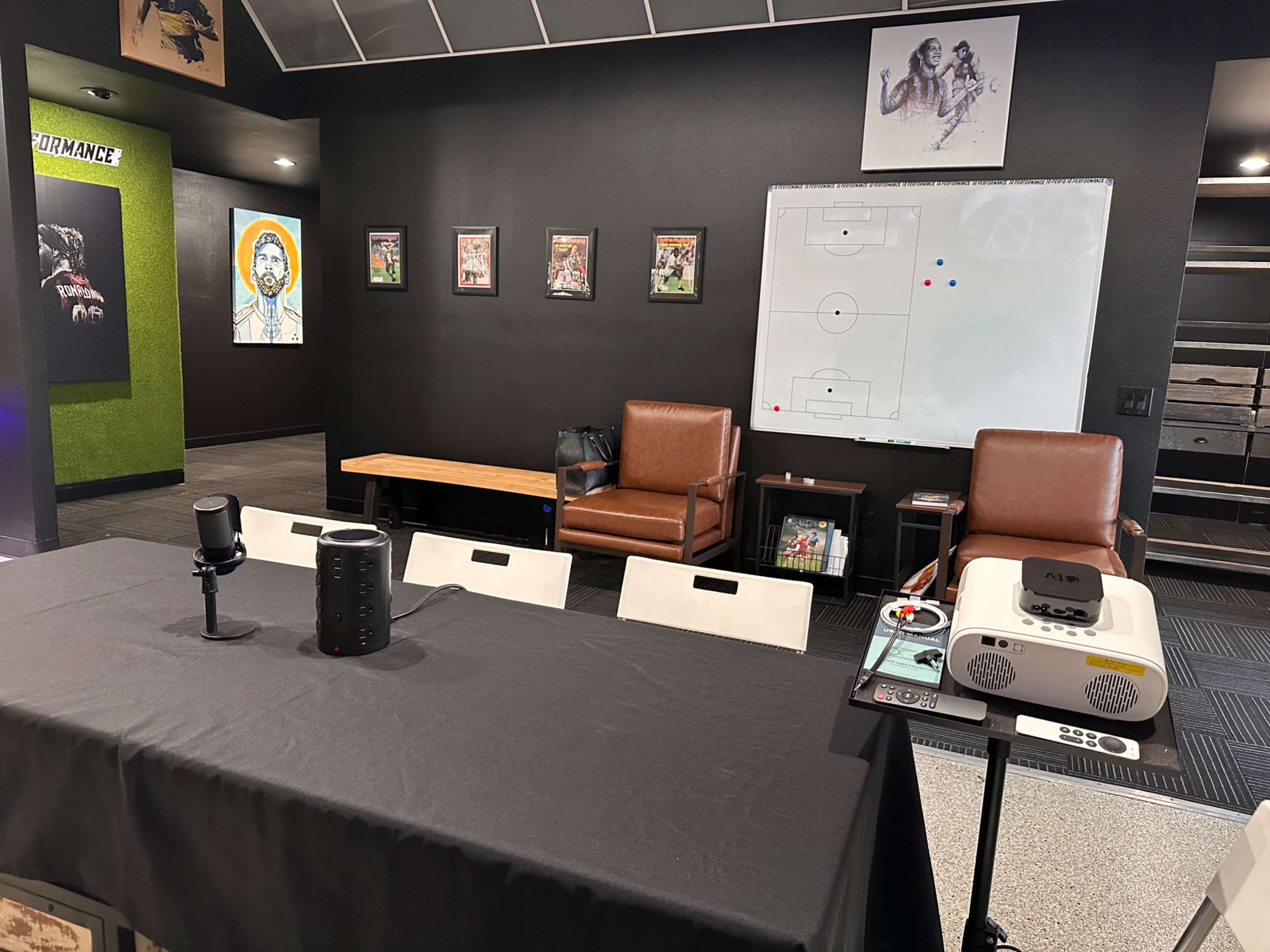 A room with a table covered by a black cloth, two brown chairs, a projector on a cart, a whiteboard with a soccer formation, and framed pictures on the walls.