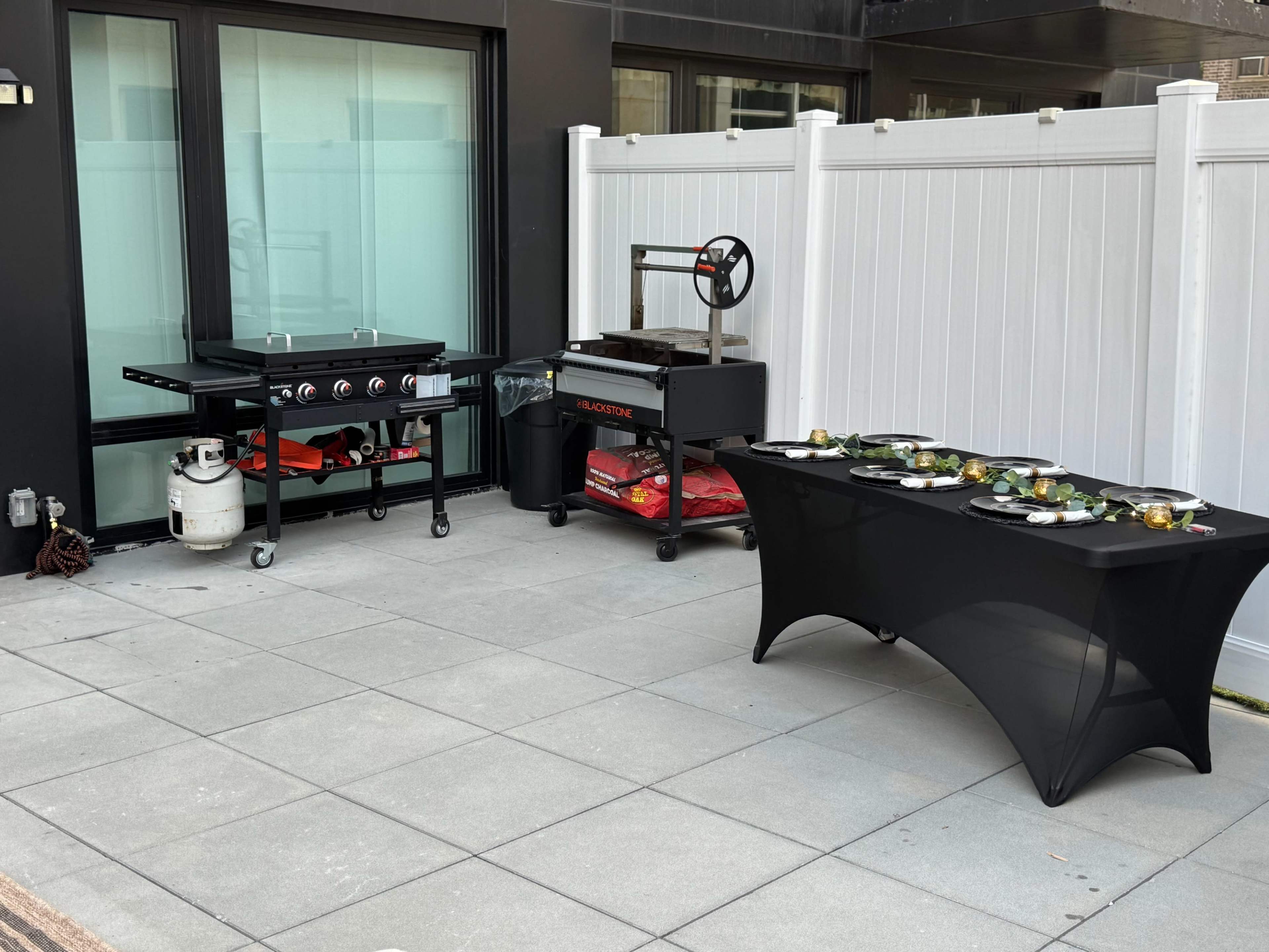 The image shows a patio area featuring a gas grill, a smoker, and a table covered with food arranged on a black tablecloth.