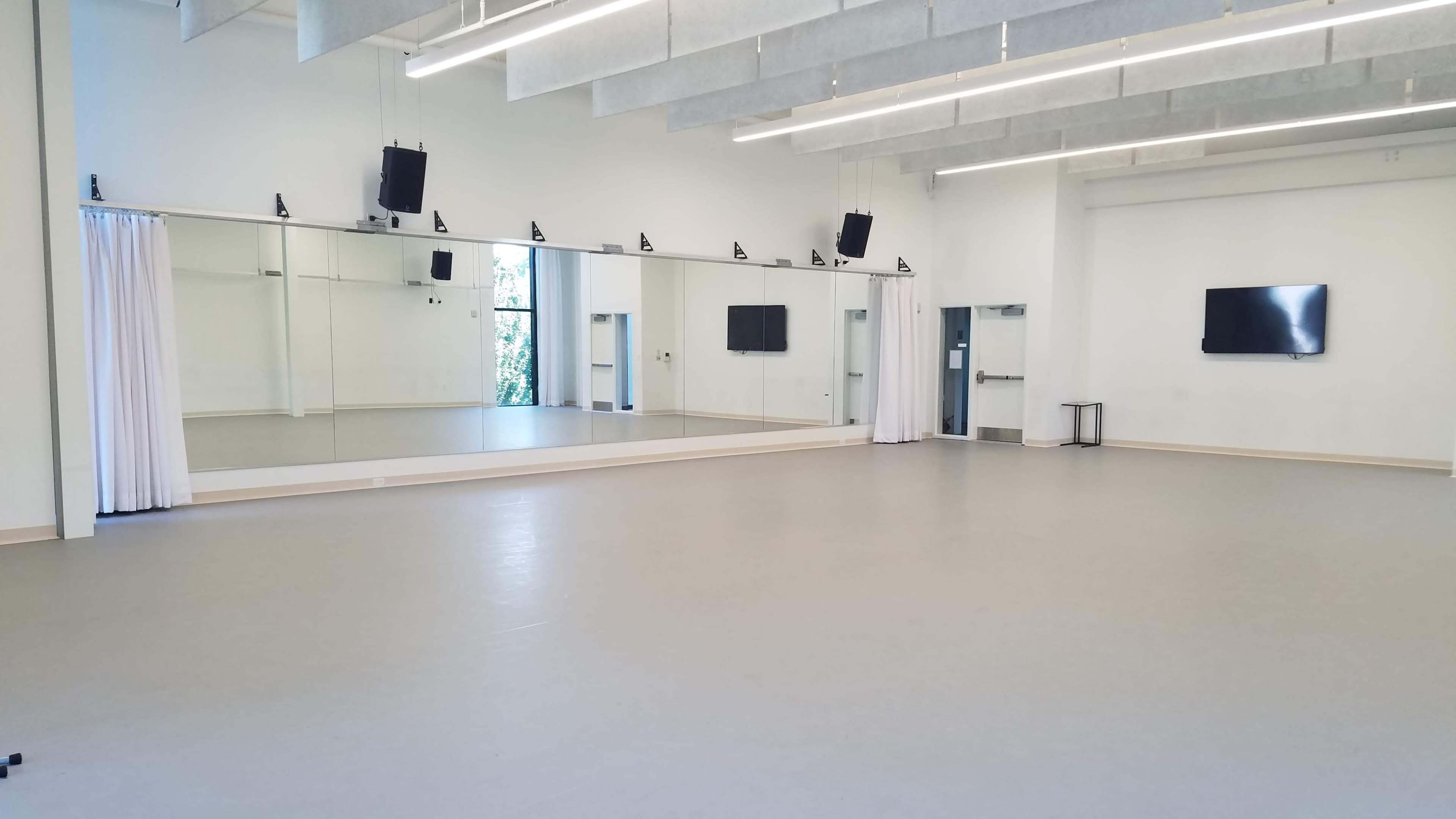 The image shows a spacious, empty dance studio with a large mirror, grey flooring, and a wall-mounted television.