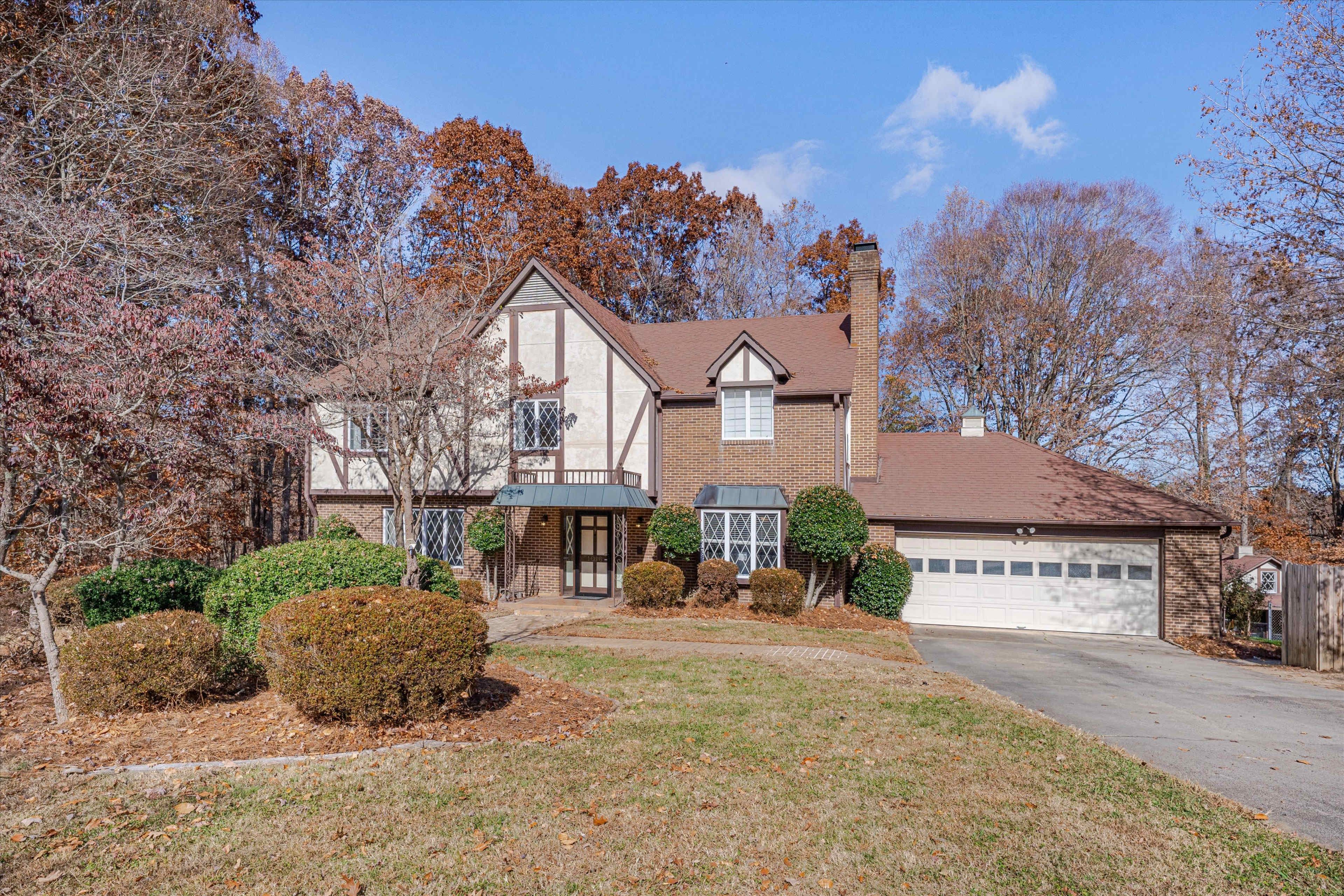 A two-story brick house with a pitched roof, a manicured front lawn, and a driveway leading to a two-car garage, surrounded by autumn-colored trees.
