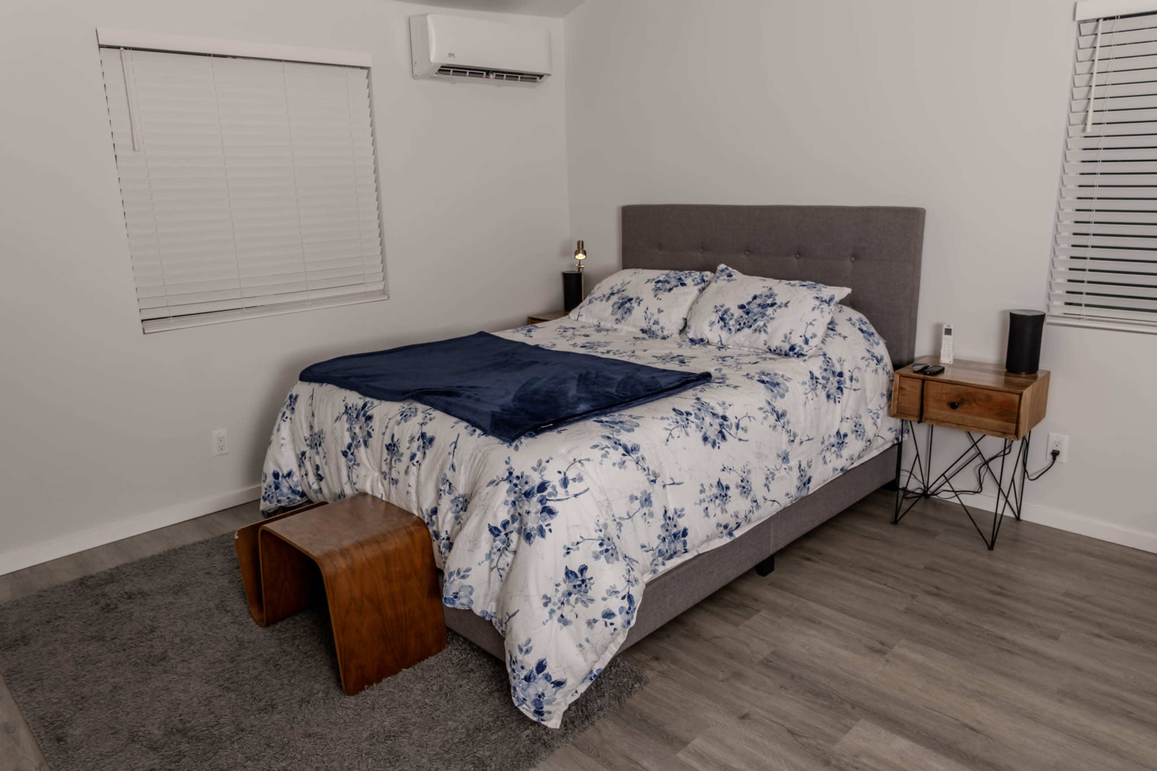 The image shows a neatly arranged bedroom featuring a bed with a floral-patterned comforter, a wooden nightstand, and a small stool on a gray rug.