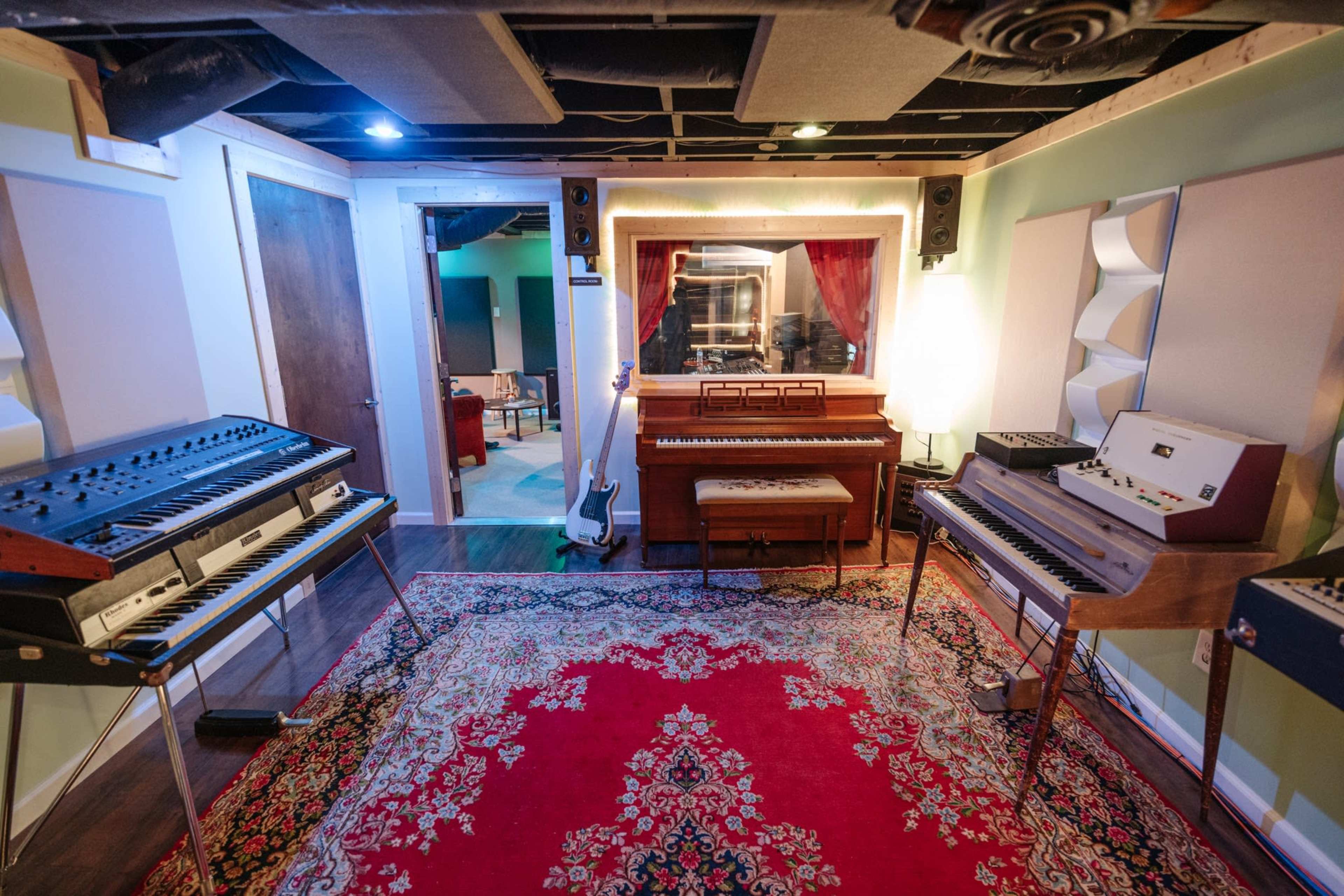 A music studio with synthesizers and a piano arranged around a central area featuring a large red area rug.