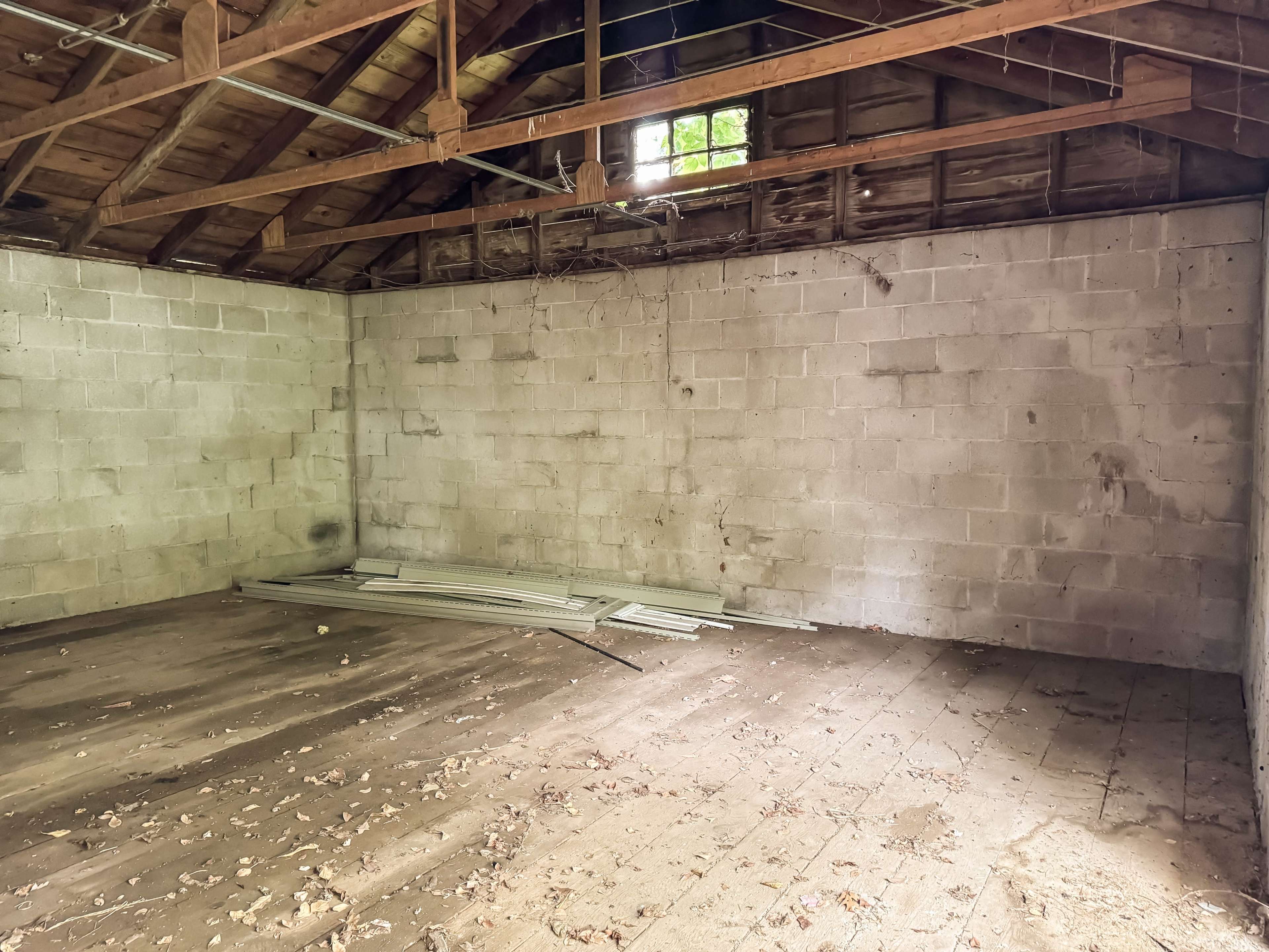 An empty, dimly lit room with exposed wooden beams, cinder block walls, and debris scattered on the wooden floor.