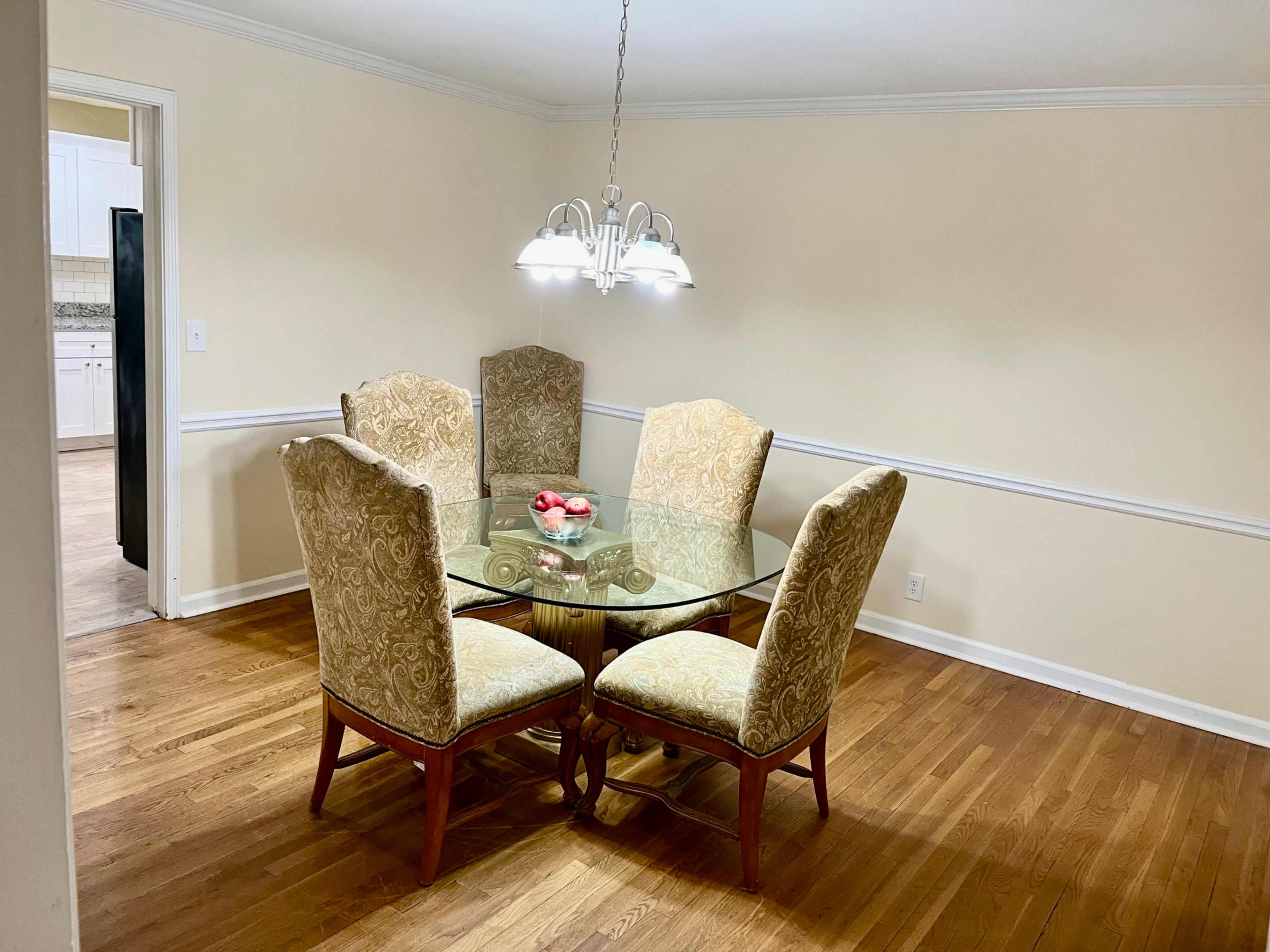A round glass dining table is surrounded by four patterned upholstered chairs in a well-lit room with hardwood flooring.