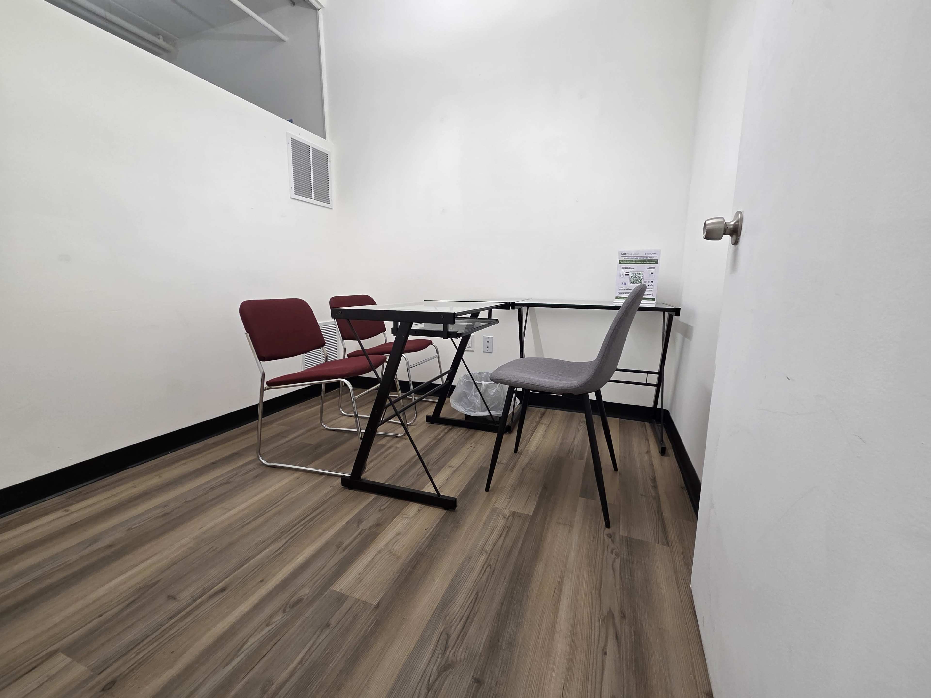 The image shows a small, empty room with two chairs and a table arranged against one wall, featuring wooden flooring and a white wall.