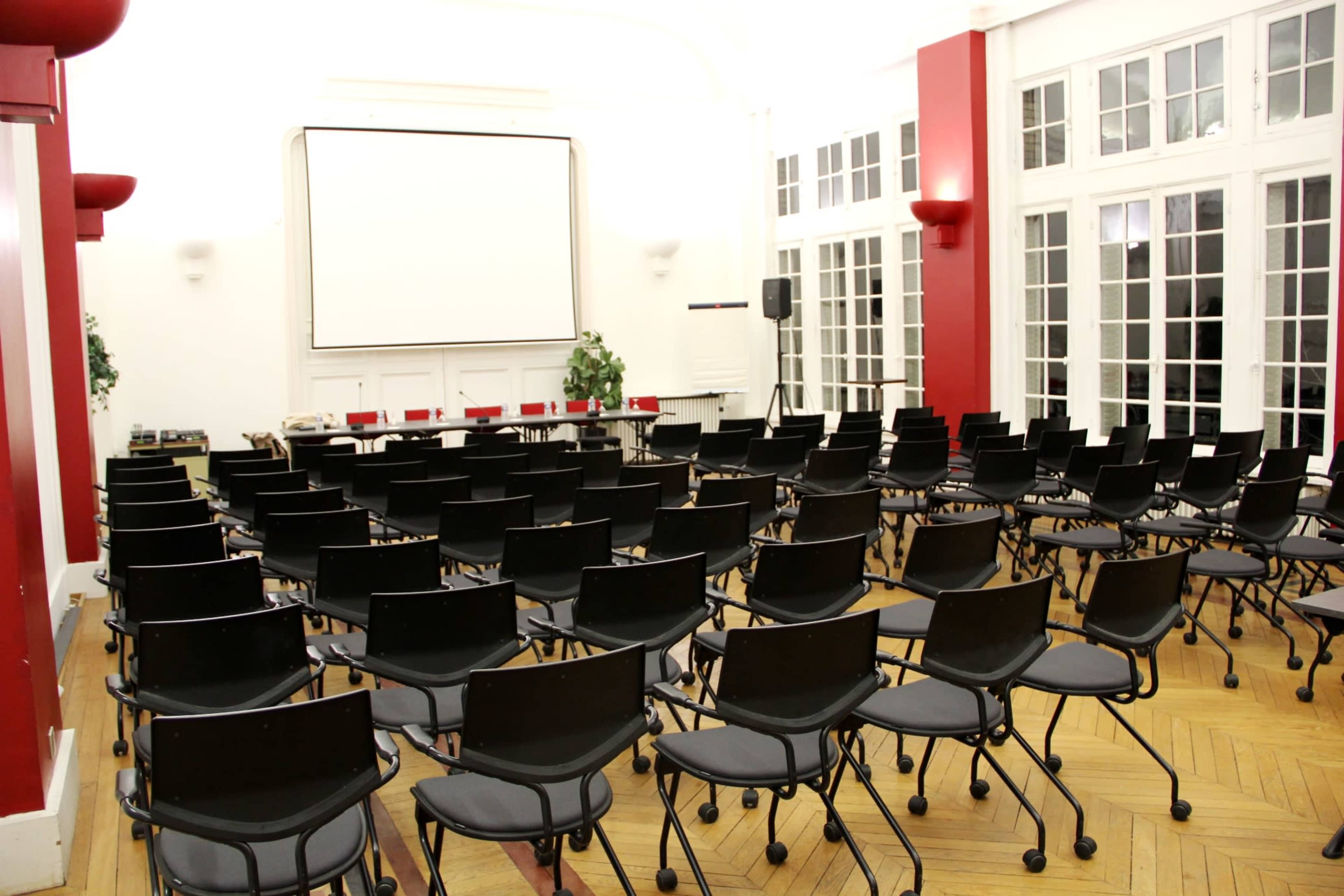 A spacious conference room set with rows of black chairs facing a blank projector screen against a bright, airy backdrop.