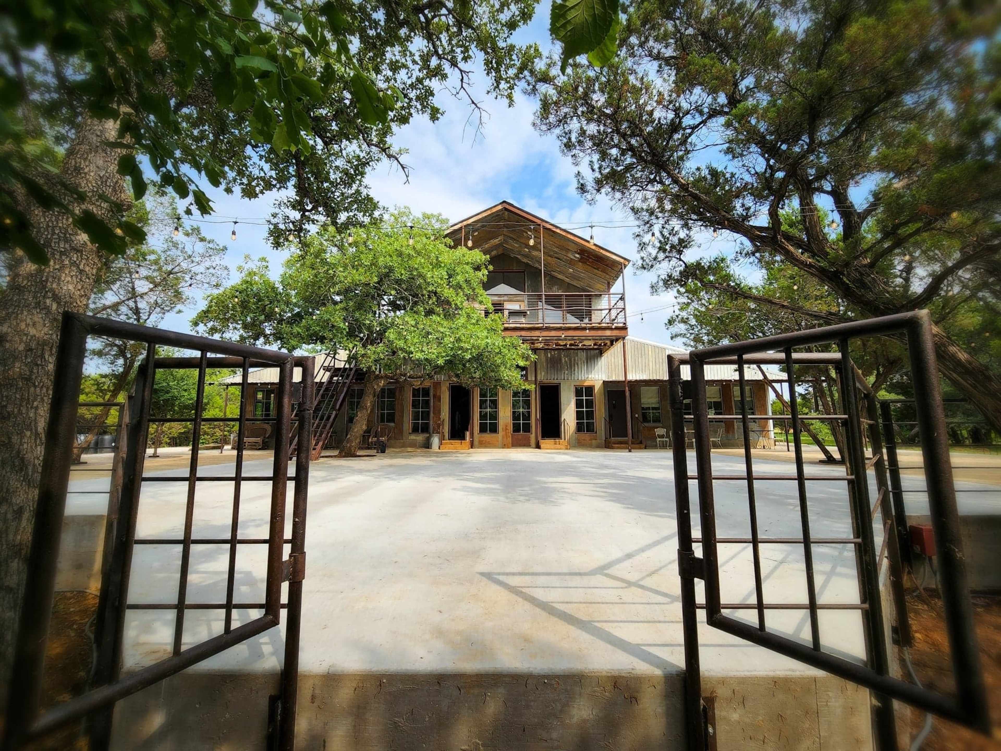The image shows a large wooden building with a balcony, surrounded by trees and a paved area, viewed through a pair of open metal gates.