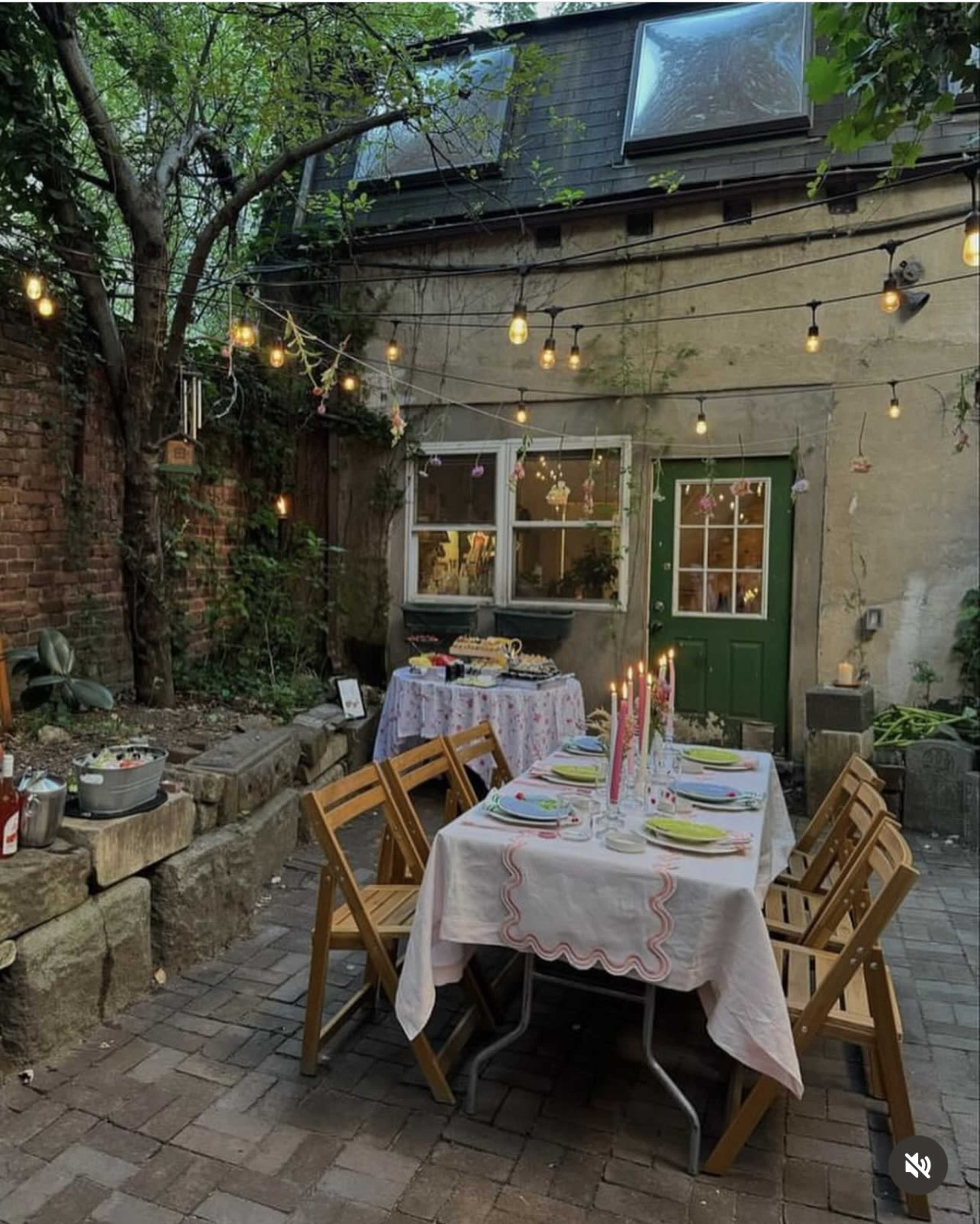 A warmly lit outdoor dining setup features a long table with candles and dishes in a garden area surrounded by trees and a brick wall.