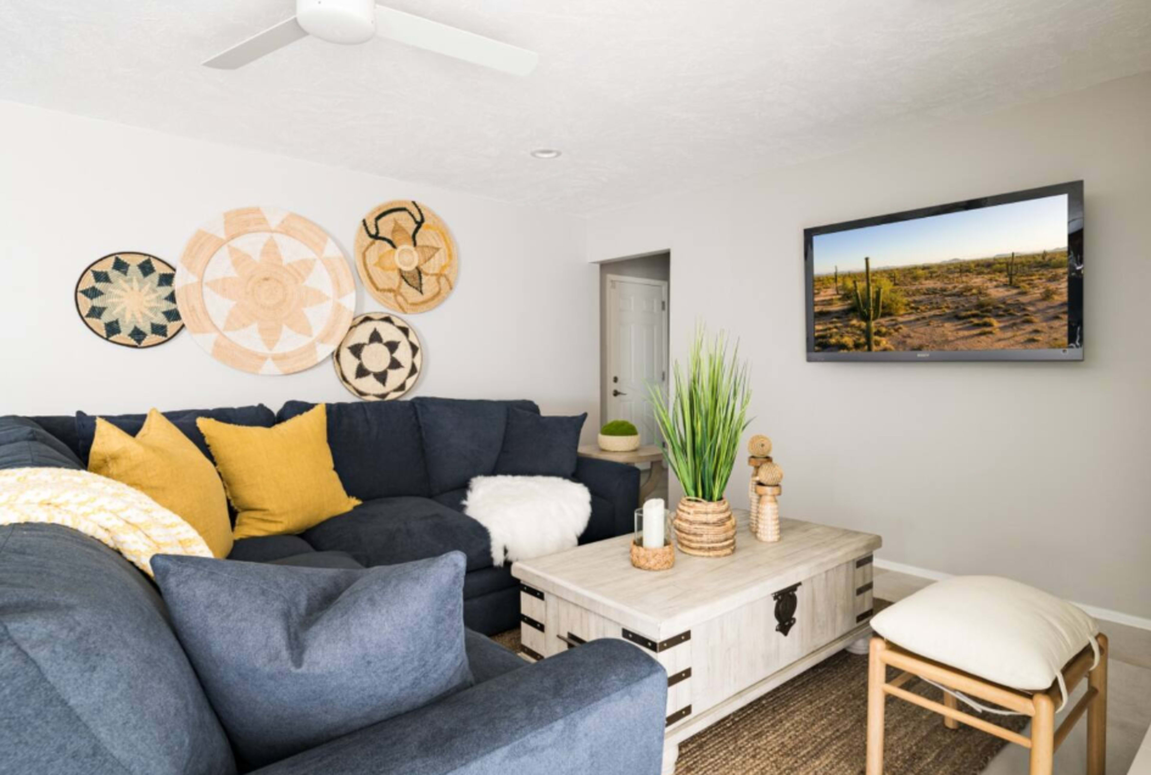 A modern living room features a dark blue sectional sofa with yellow throw pillows, a wooden coffee table, and wall decor made of woven baskets, with a television displaying a desert landscape.