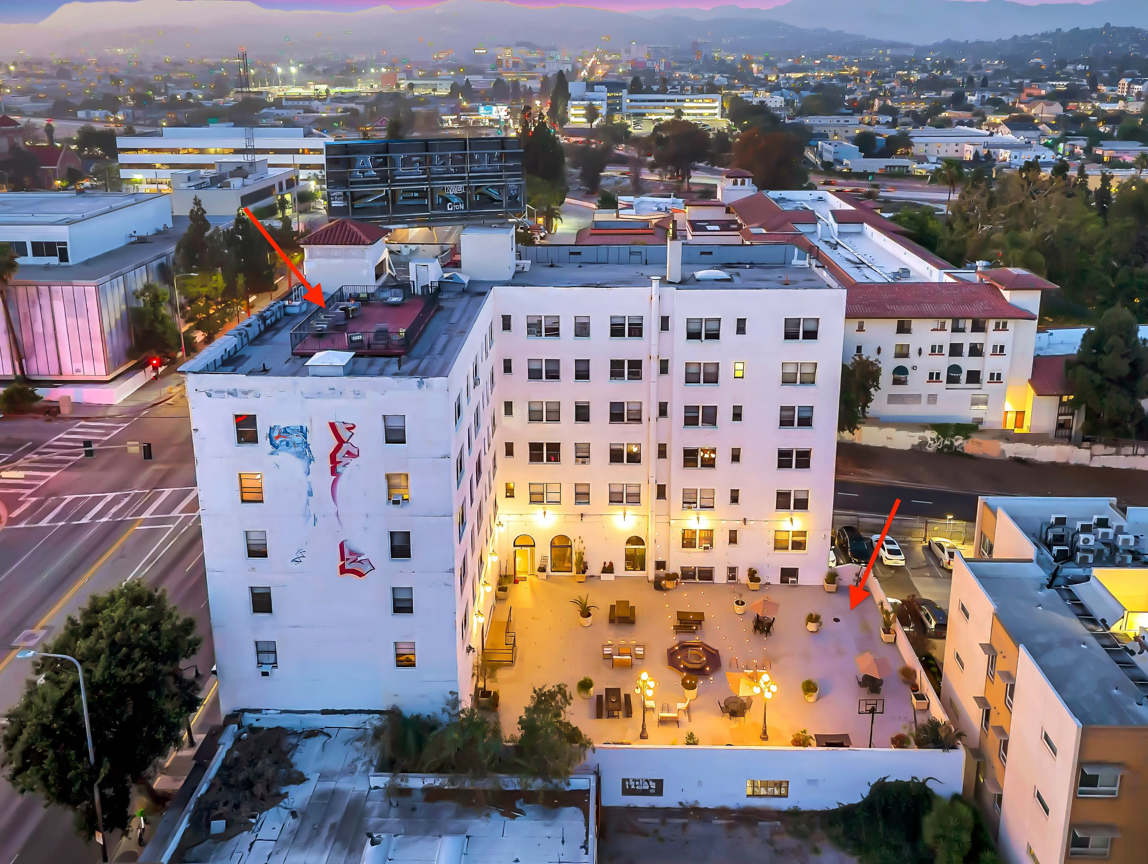 The image shows an aerial view of a multi-story white building with a courtyard and various outdoor seating arrangements, surrounded by urban structures and roads.