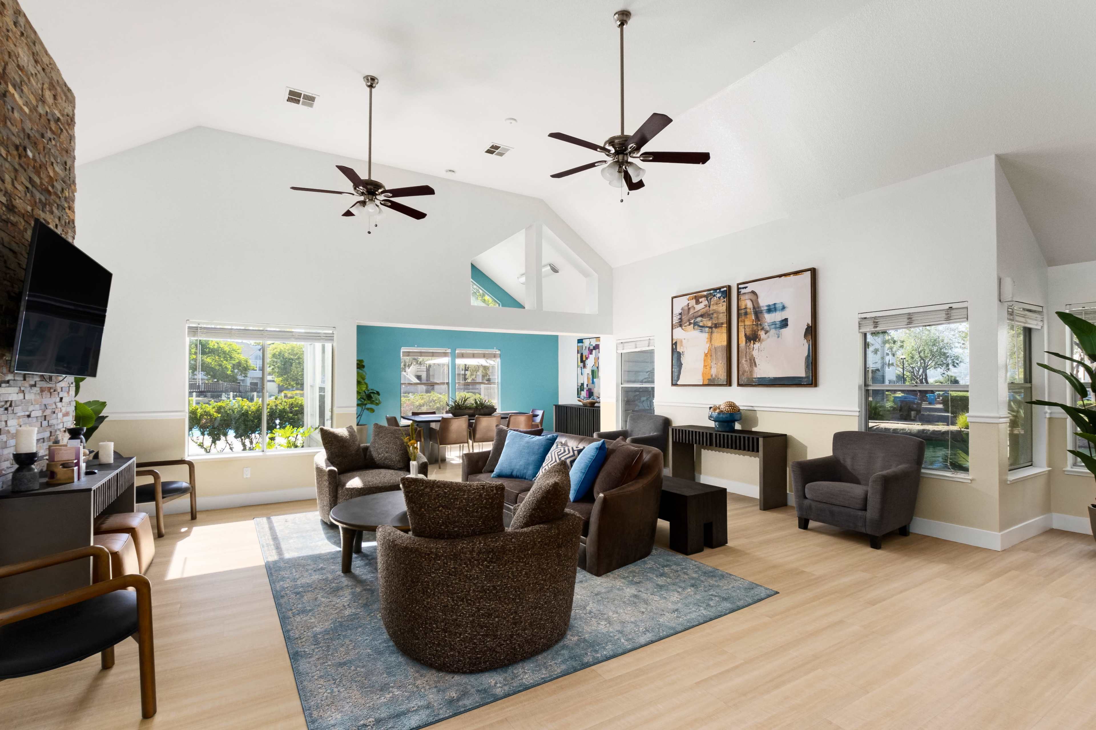 The image shows a modern living space with a high ceiling, a stone accent wall, comfortable seating areas, and large windows allowing natural light to enter.