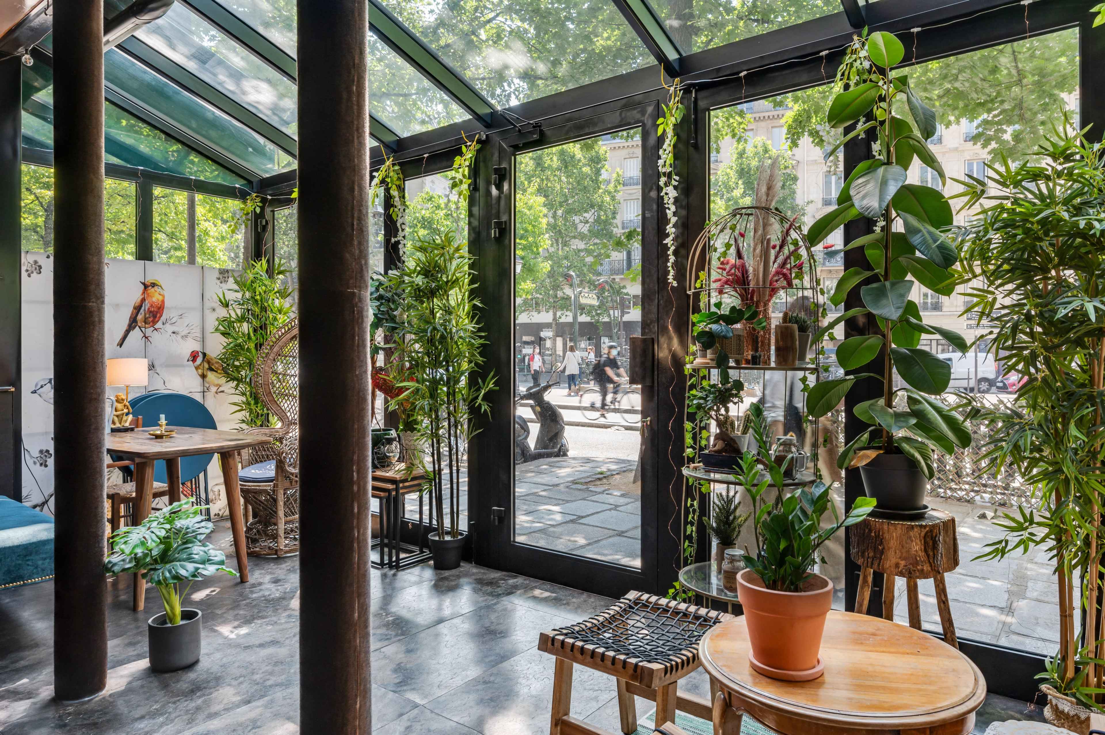 A glass-walled indoor space filled with plants, featuring wooden tables and chairs, overlooks a street with people walking by.